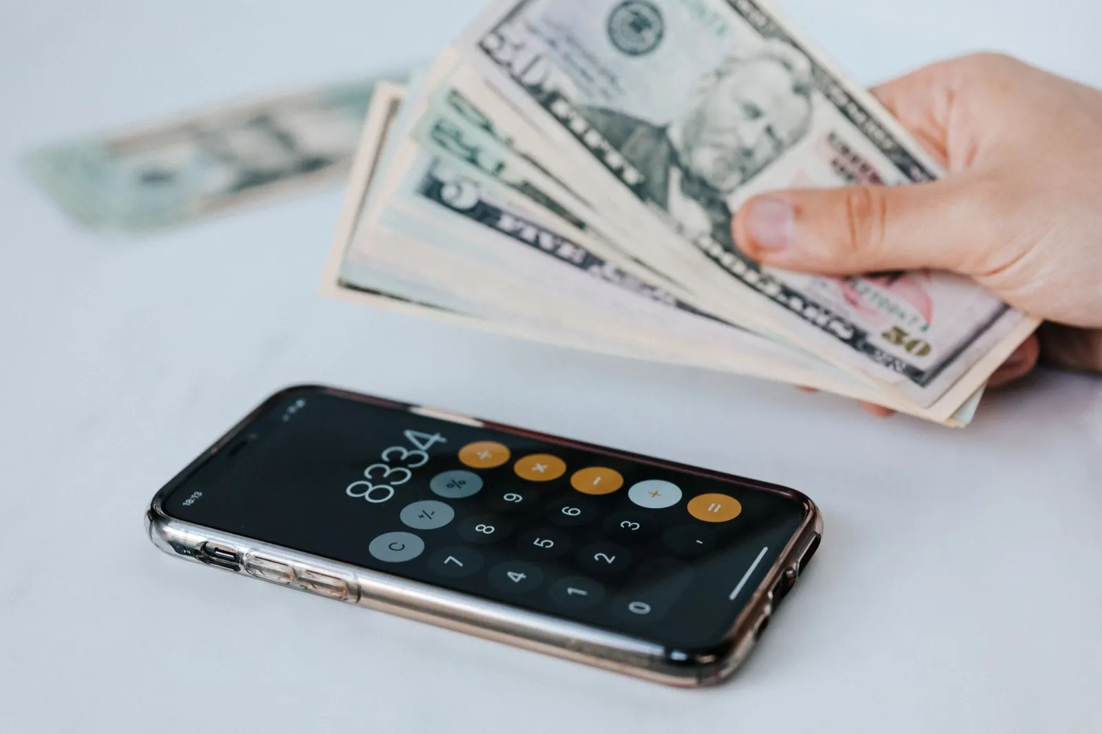 Calculator application on mobile phone screen on white surface near crop anonymous person holding pile of dollars. Stock photo illustrating Emerald Finance's partnership story.