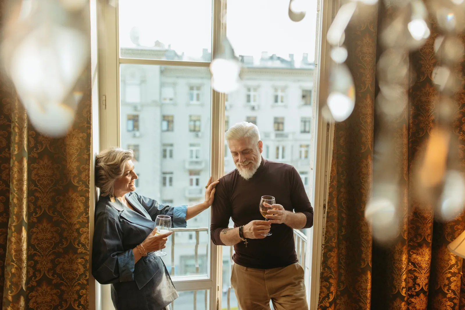 A senior couple enjoys wine and laughter by balcony windows inside a cozy apartment. Stock photo illustrating Max India's earning call story.