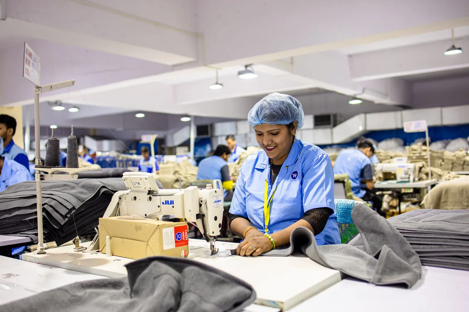 Smiling factory workers operate sewing machines in a bustling textile factory environment. Stock photo illustrating AYM Syntex's financial results story.