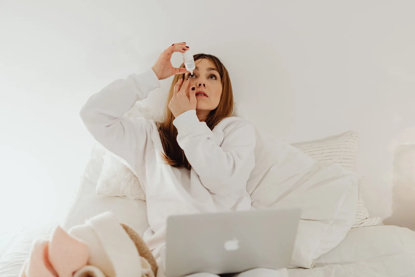 A woman applying eye drops while resting in bed with a laptop, symbolizing relaxation and self-care at home. Stock photo illustrating Lupin's product launched story.