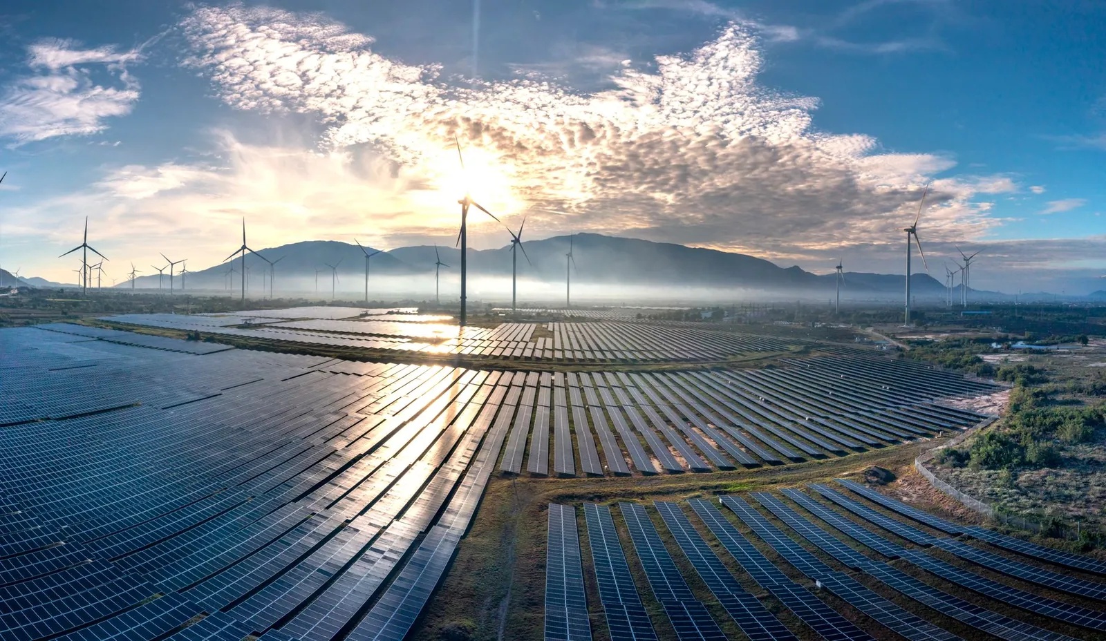 Stunning aerial view of a vast solar farm with wind turbines under a vibrant sunrise sky, showcasing renewable energy. Stock photo illustrating NTPC Green Energy's production started story.