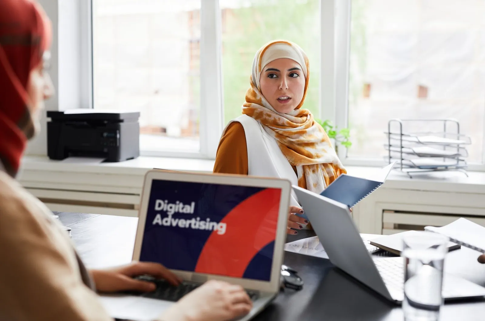 Two women in hijabs discuss digital advertising in an office setting. Stock photo illustrating Mobavenue AI Tech's product launched story.