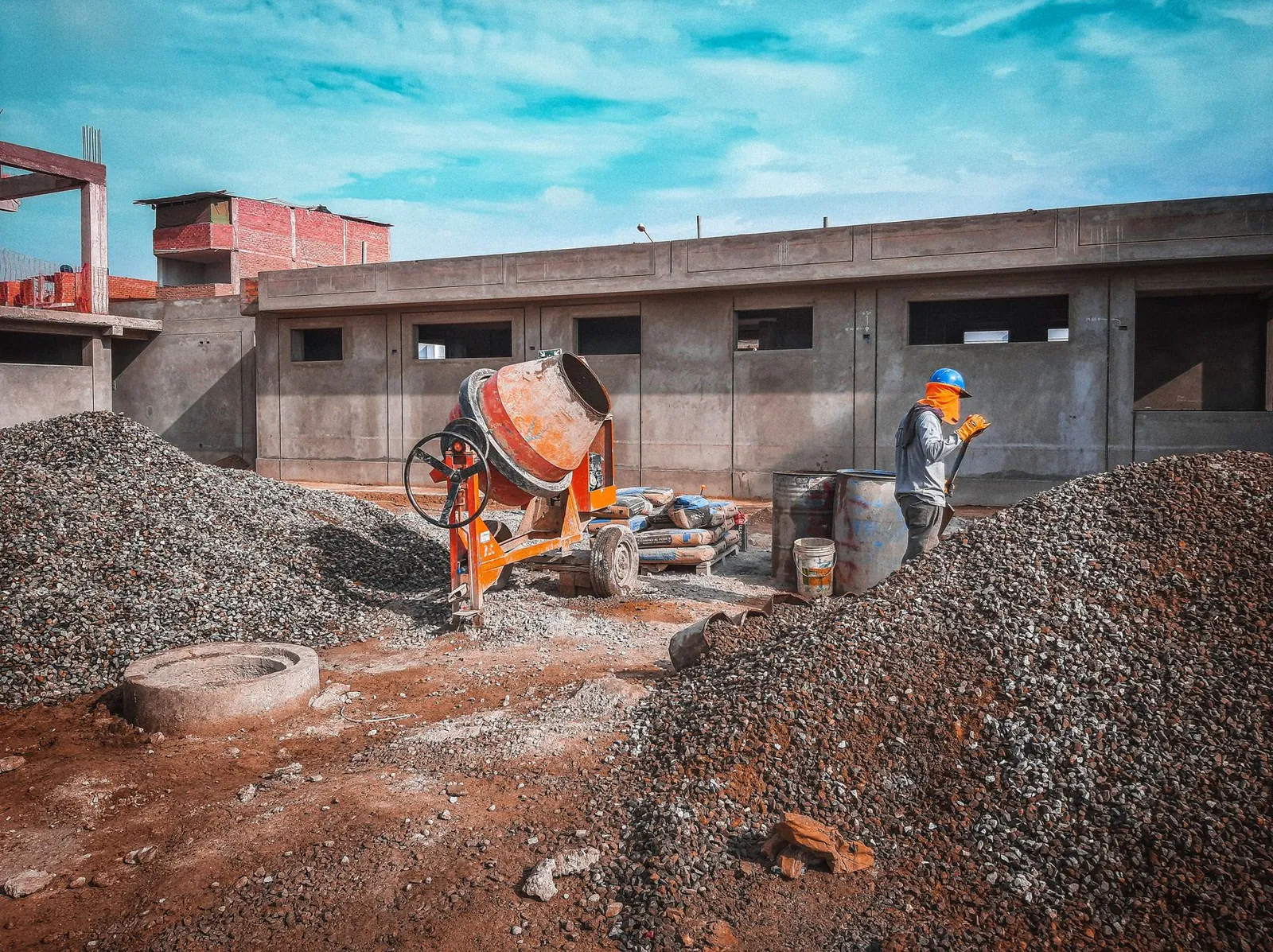 A construction site with a worker wearing safety gear next to a cement mixer and gravel piles. Stock photo illustrating Prism Johnson's fund raise story.