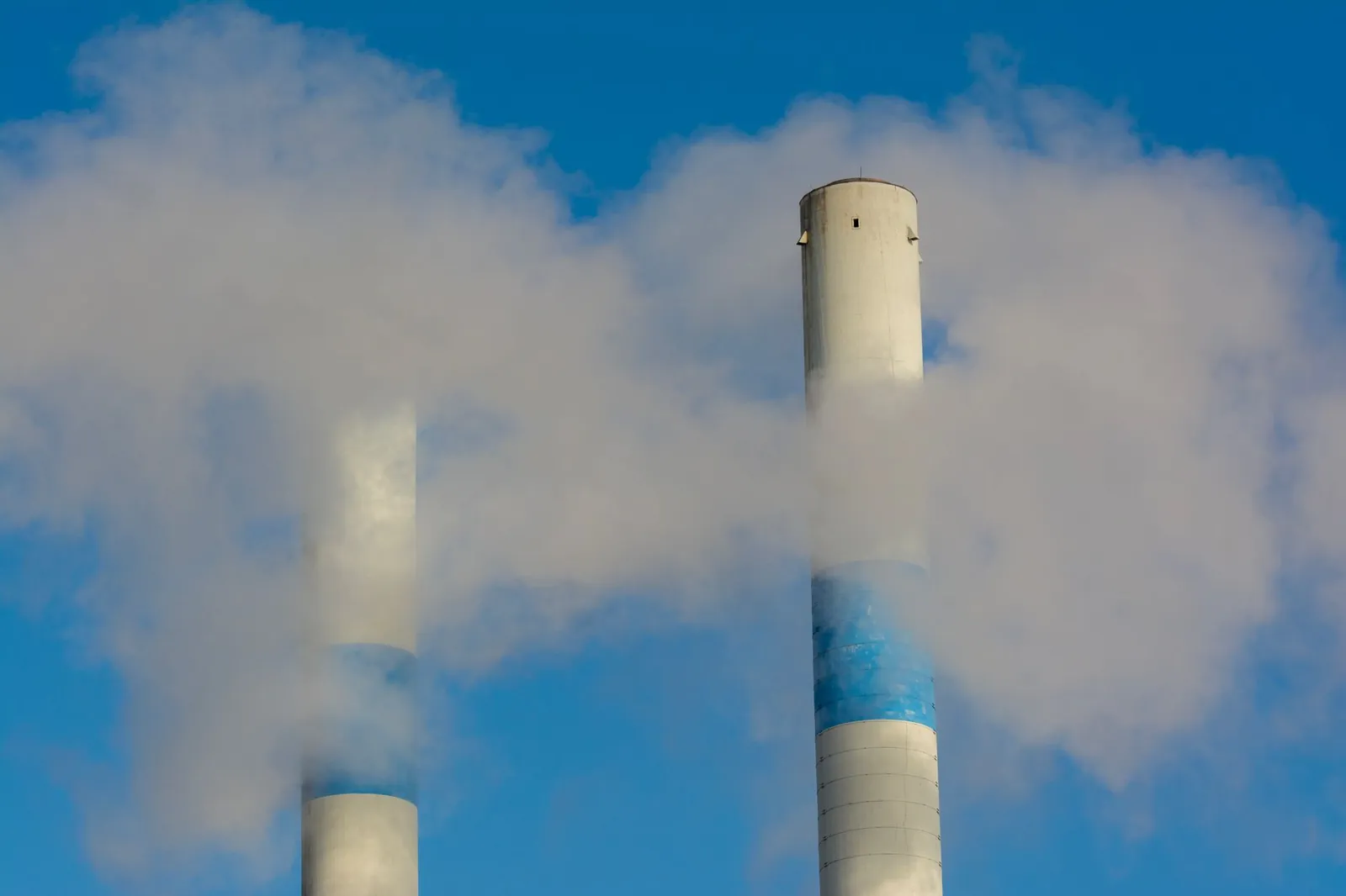 Twin industrial chimneys emitting smoke under a clear blue sky, highlighting air pollution concerns. Stock photo illustrating Linde India's production started story.