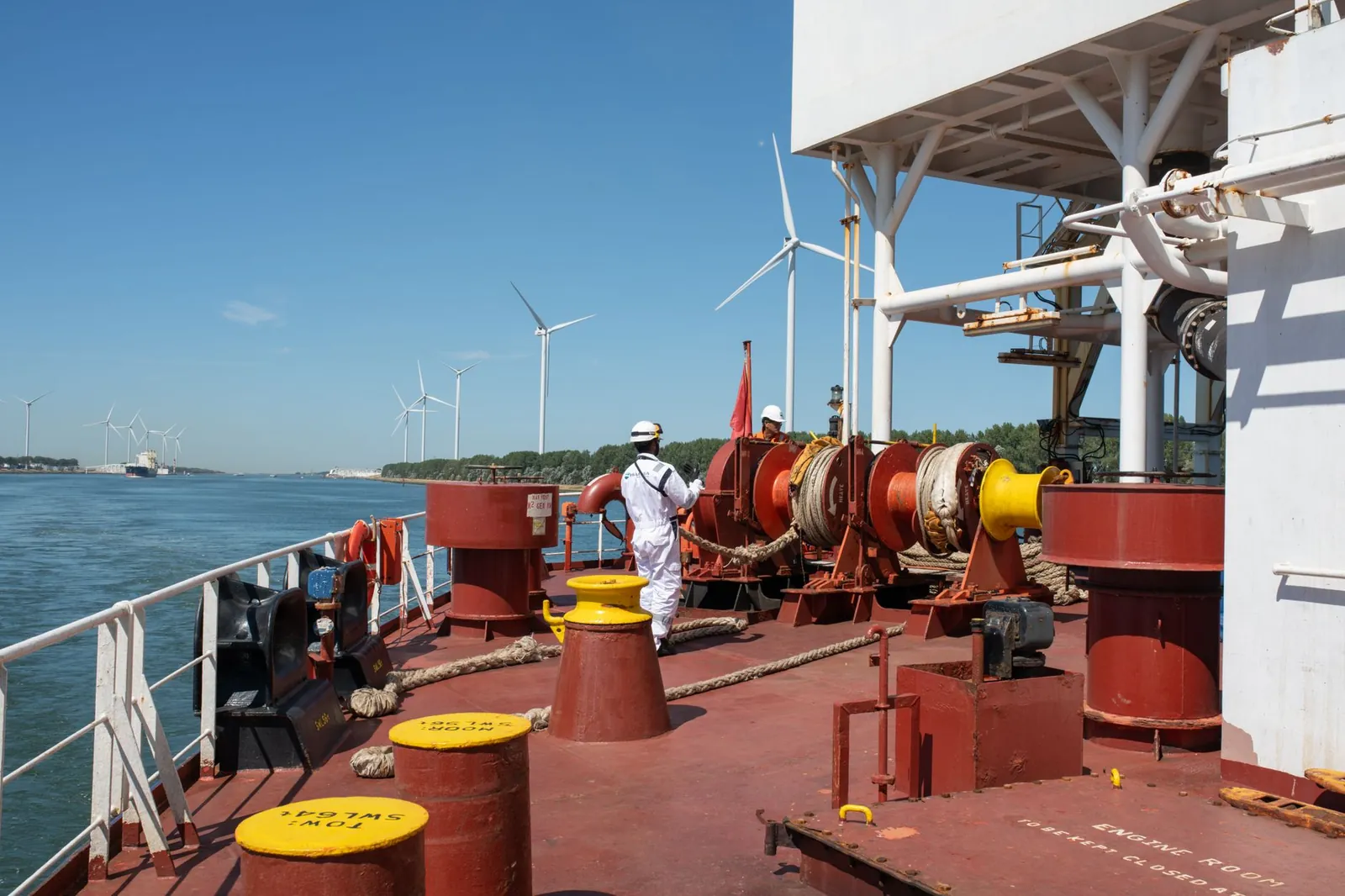 Sailor managing equipment on a ship with offshore wind turbines in the background under a clear blue sky. Stock photo illustrating Inox Green Energy Services' fund raise story.