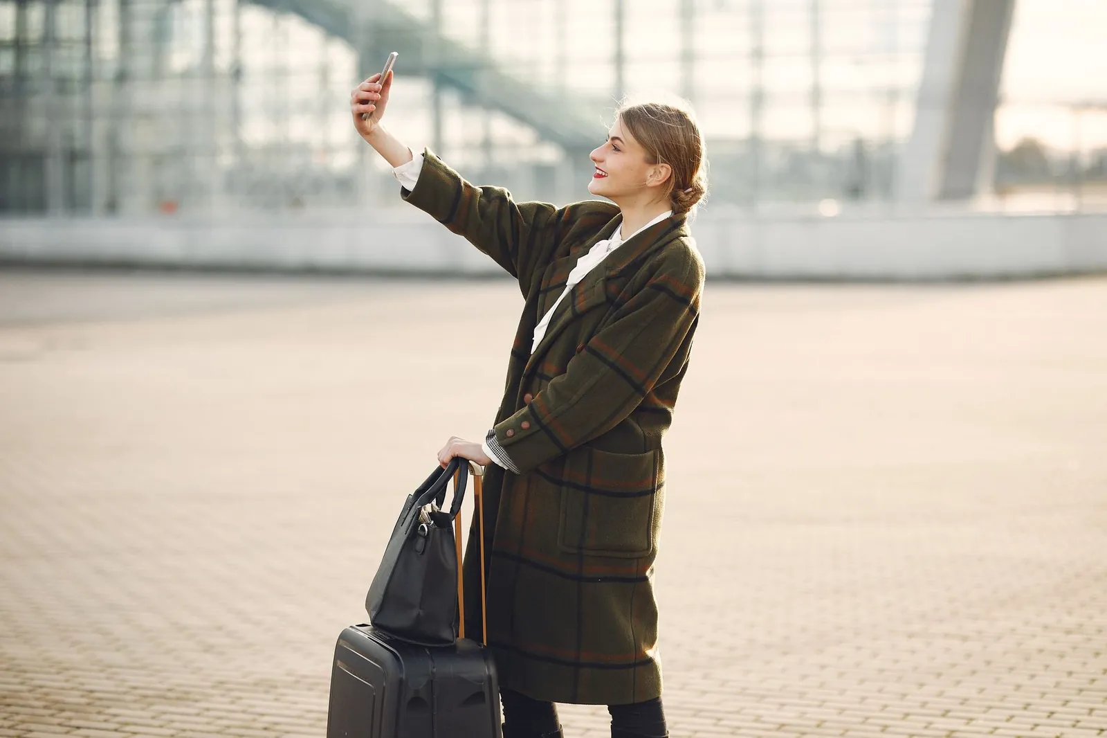 Side view of smiling cheerful female traveler with suitcase and bag wearing trendy plaid coat using smartphone for selfie while standing against contemporary building on city street. Stock photo illustrating Tata Communications' product launched stor