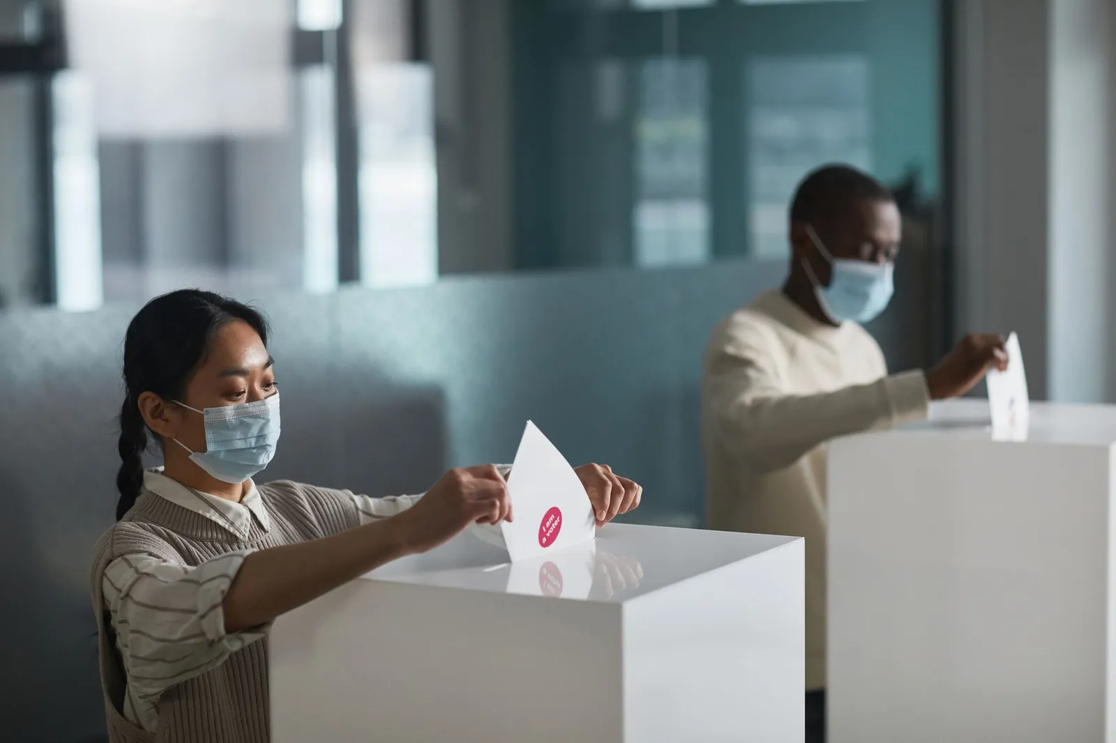 People casting ballots in an indoor election setting, practicing safety measures with masks. Stock photo for the ALFA News guide: Understanding the Postal Ballot Notice in the Indian Stock Market.