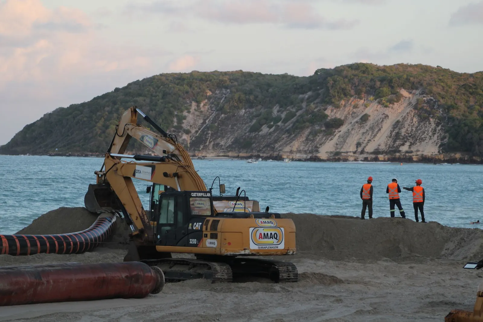 Construction workers and equipment on a beach with hills and ocean backdrop. Stock photo illustrating Dilip Buildcon's new order story.