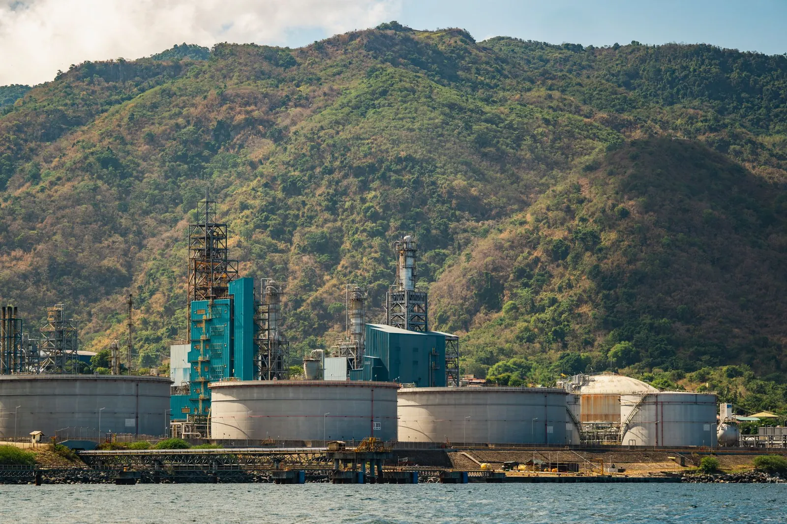 Industrial plant with large tanks and chimneys against a lush mountain backdrop. Stock photo illustrating SPML Infra's new order story.