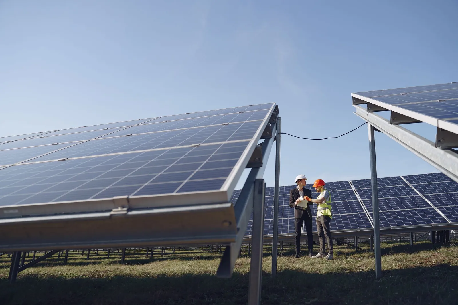 Supervisor in hardhat and formal wear discussing project with workman in uniform while standing with papers near modern solar panels under blue sky. Stock photo illustrating NTPC's production started story.