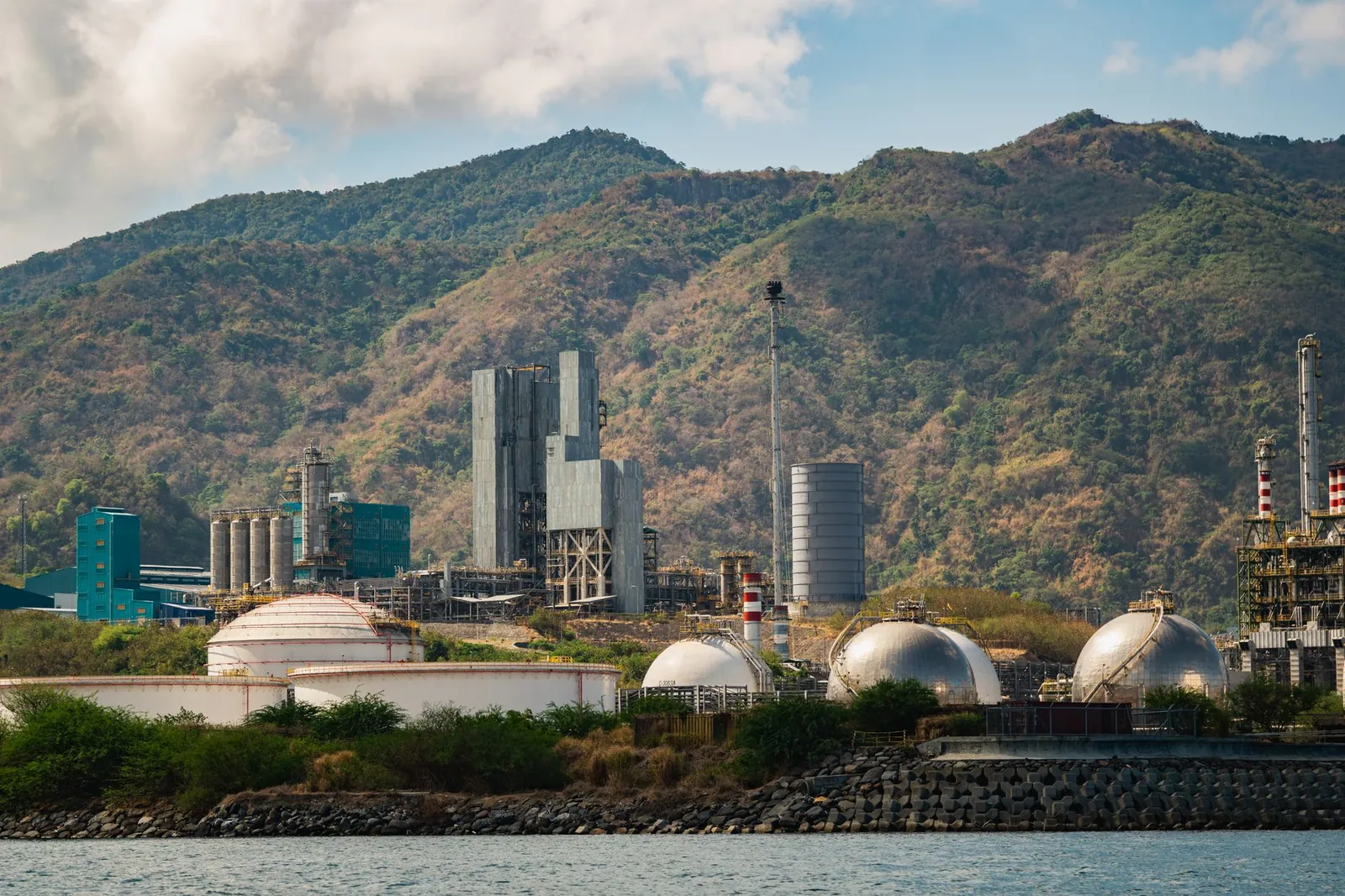 Industrial gas tanks and chimneys against lush hills, showcasing industry and nature. Stock photo illustrating Chemplast Sanmar's production started story.