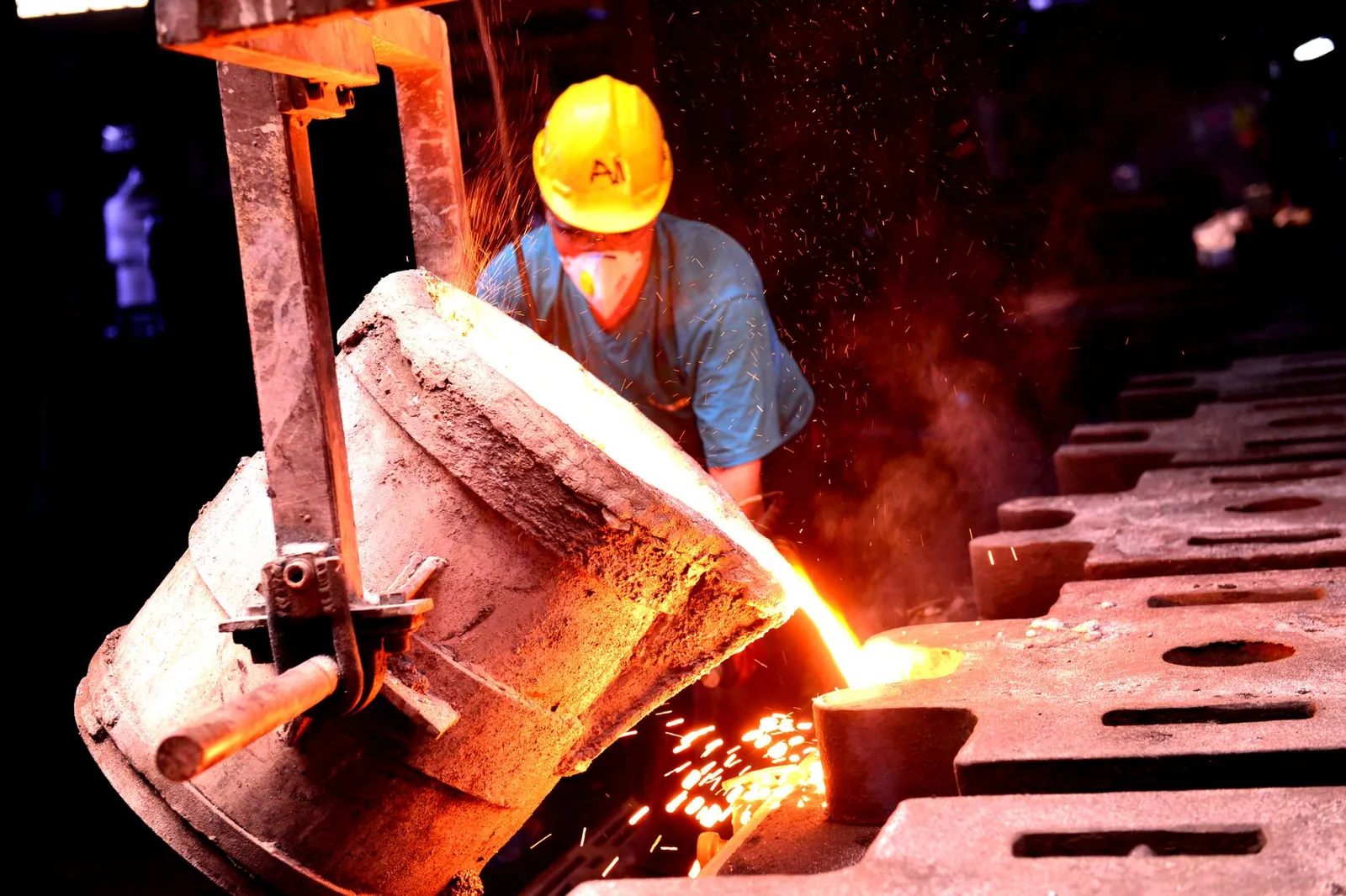 A factory worker wearing a helmet pours molten metal into molds, showcasing industrial metal casting. Stock photo illustrating Simplex Castings' fund raise story.