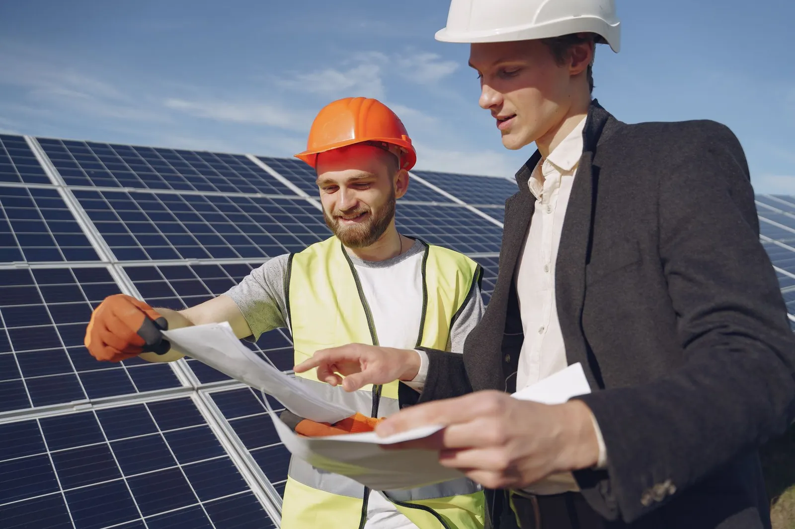 Low angle of young engineer and businessman in hardhats discussing project details against solar energy station. Stock photo illustrating Clean Max Enviro Energy Solutions' production started story.