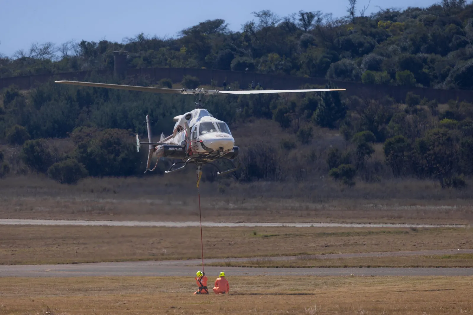 A helicopter hovers above a grassy field with two crew members on the ground in safety gear. Stock photo illustrating Flywings Simulator Training Centre's regulatory approval story.