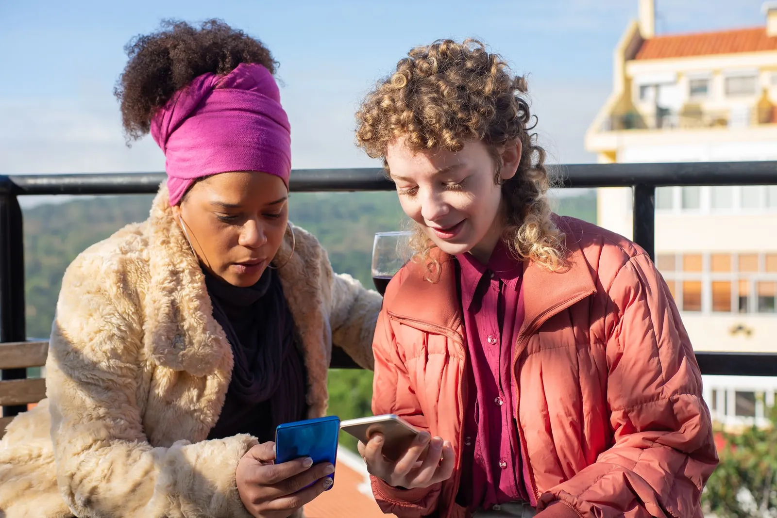 Two women sitting on a rooftop, enjoying wine and using smartphones together. Stock photo illustrating One 97 Communications' product launched story.