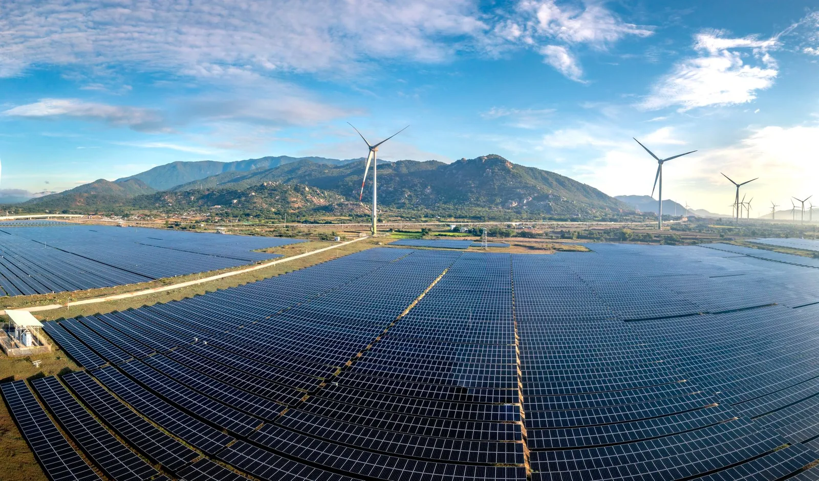 Aerial view of a vast solar farm with wind turbines against a mountainous backdrop on a sunny day. Stock photo illustrating Enviro Infra Engineers' new order story.