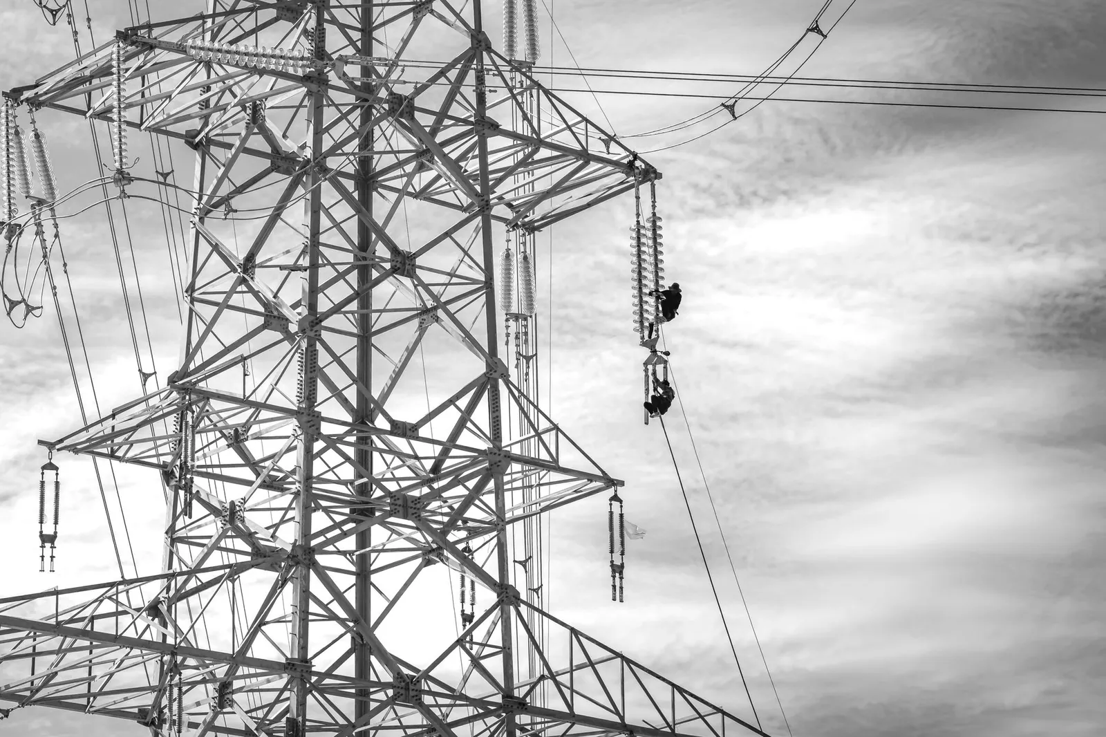 Black and white image of workers performing maintenance on a transmission tower against a cloudy sky. Stock photo illustrating Vikran Engineering's earning call story.