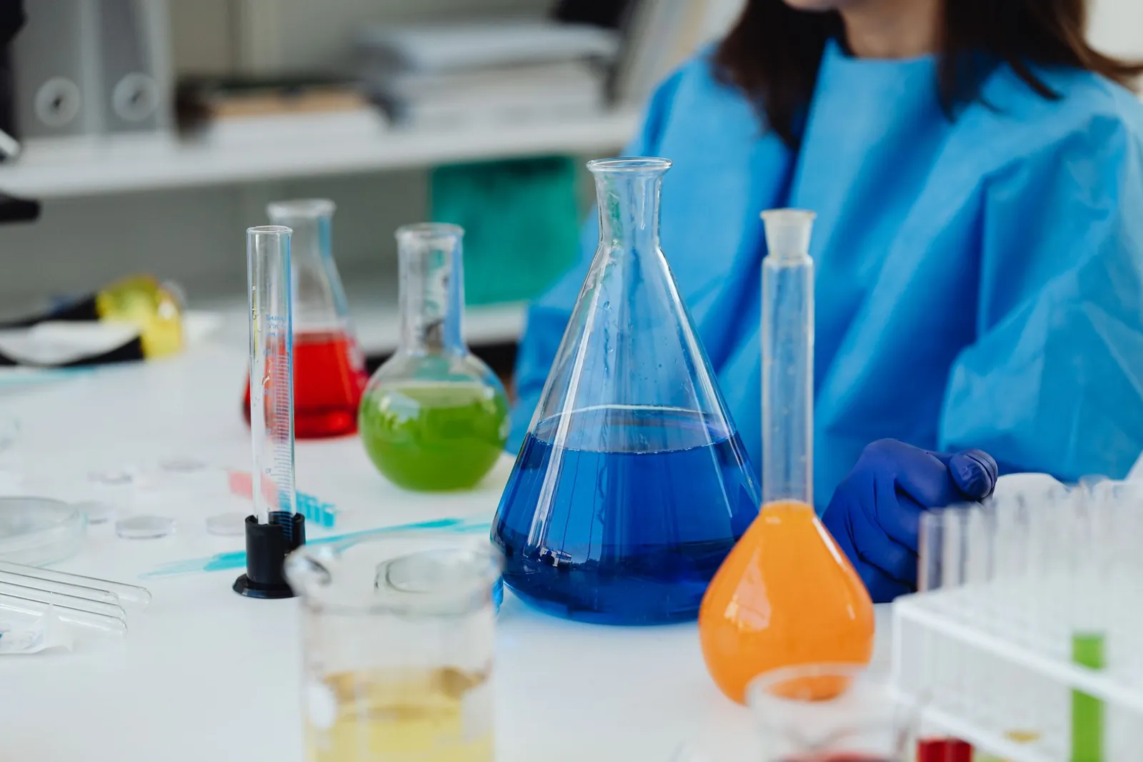 A close-up of a laboratory desk with colorful chemicals in glassware, featuring a researcher in protective gear. Stock photo illustrating India Pesticides' product launched story.