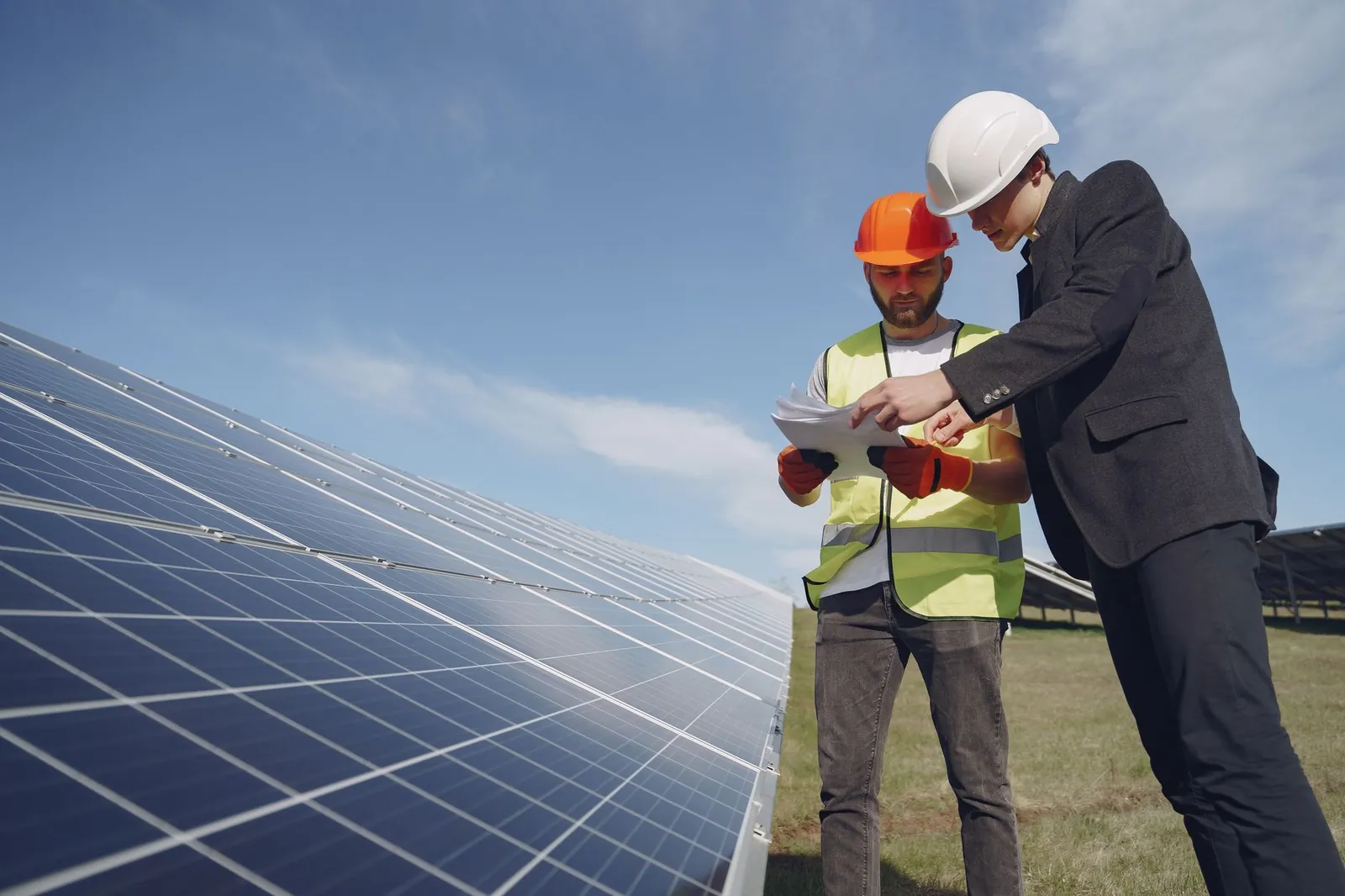 Low angle of young inspector and foreman in hardhats checking documentation against modern solar panels in field. Stock photo illustrating NTPC Green Energy's production started story.