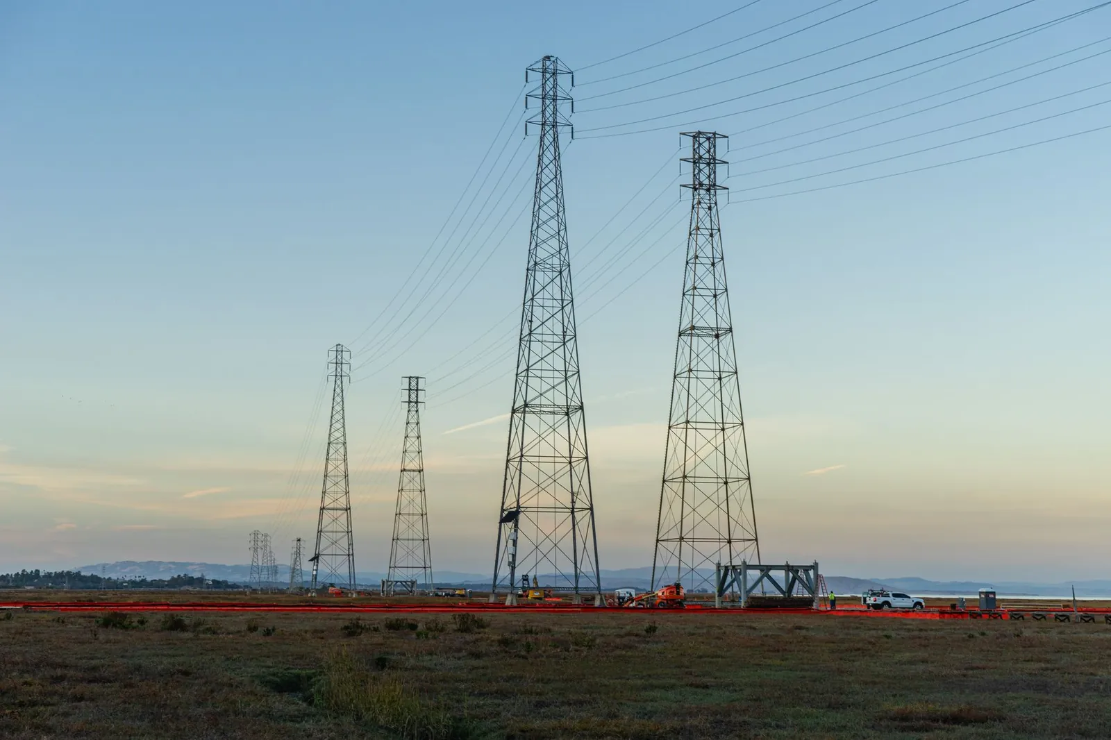 High voltage transmission towers with clear blue sky at dusk in an open field. Stock photo illustrating Dilip Buildcon's new order story.