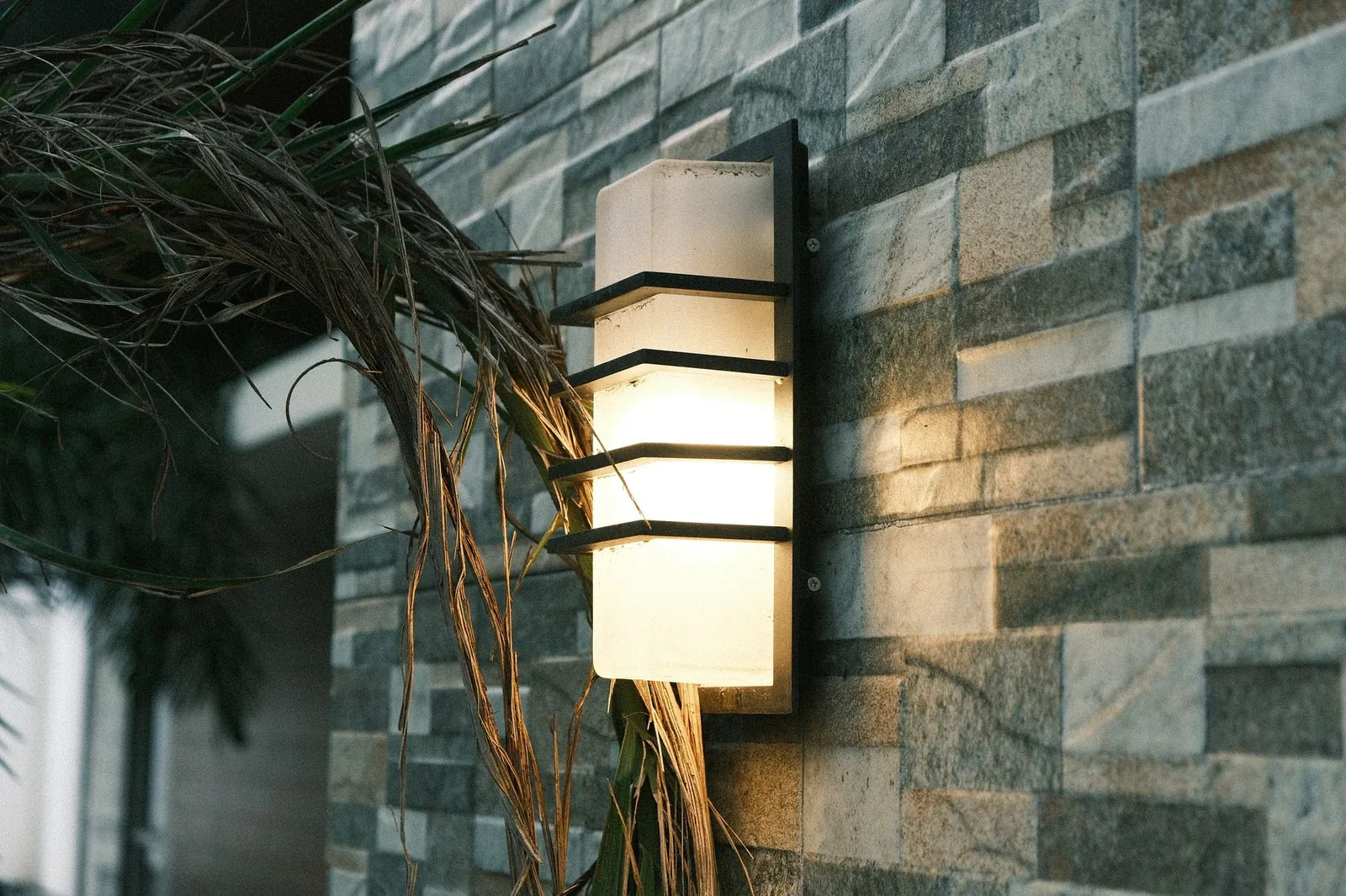Illuminated outdoor wall lantern on a textured stone wall, surrounded by plants, setting a cozy evening ambiance. Stock photo illustrating Crompton Greaves Consumer Electricals' product launched story.