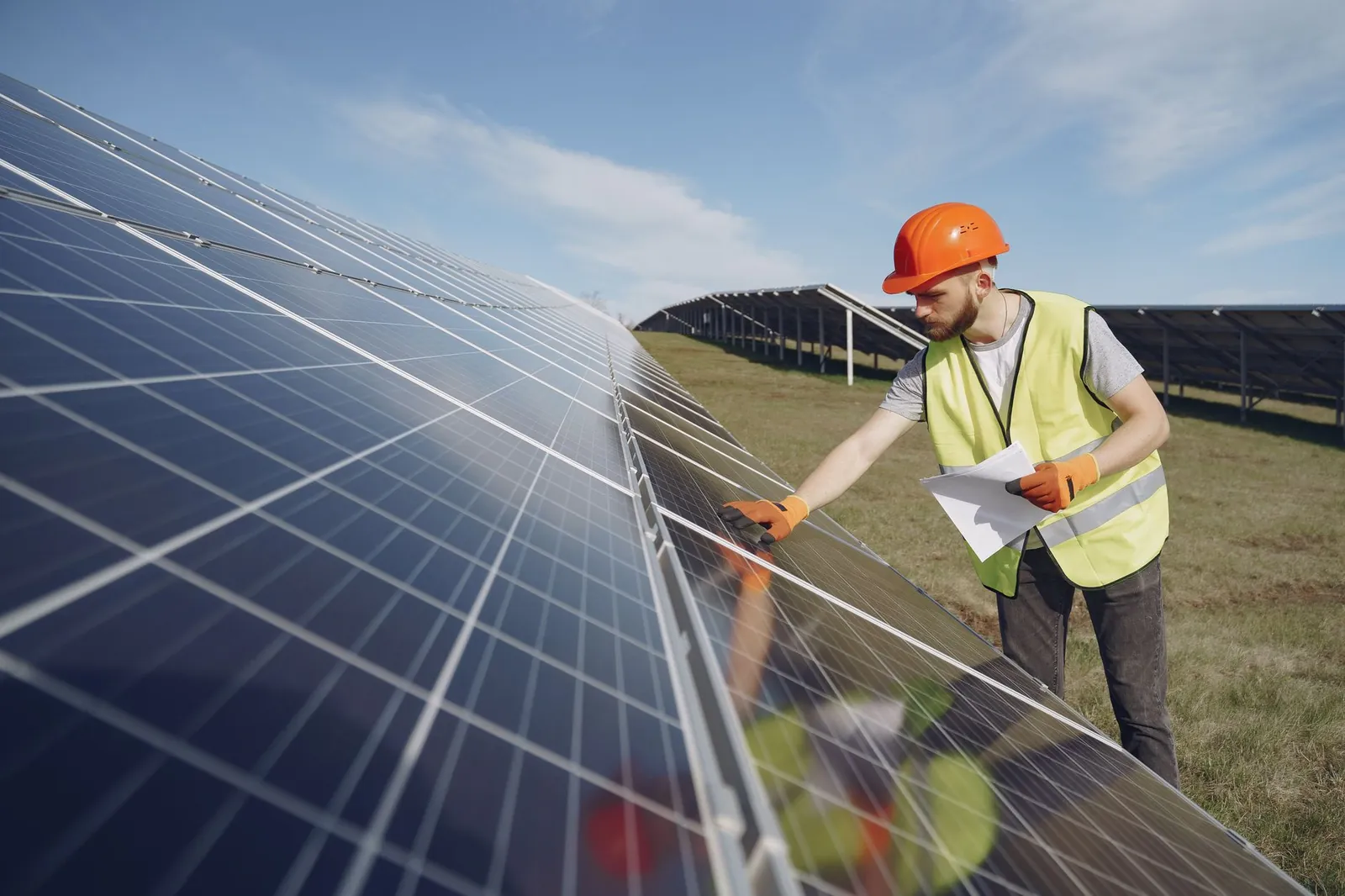 Young bearded male engineer in protective helmet and heat resistant gloves touching solar panel while standing on field with papers on sunny day. Stock photo illustrating Bluspring Enterprises' new order story.