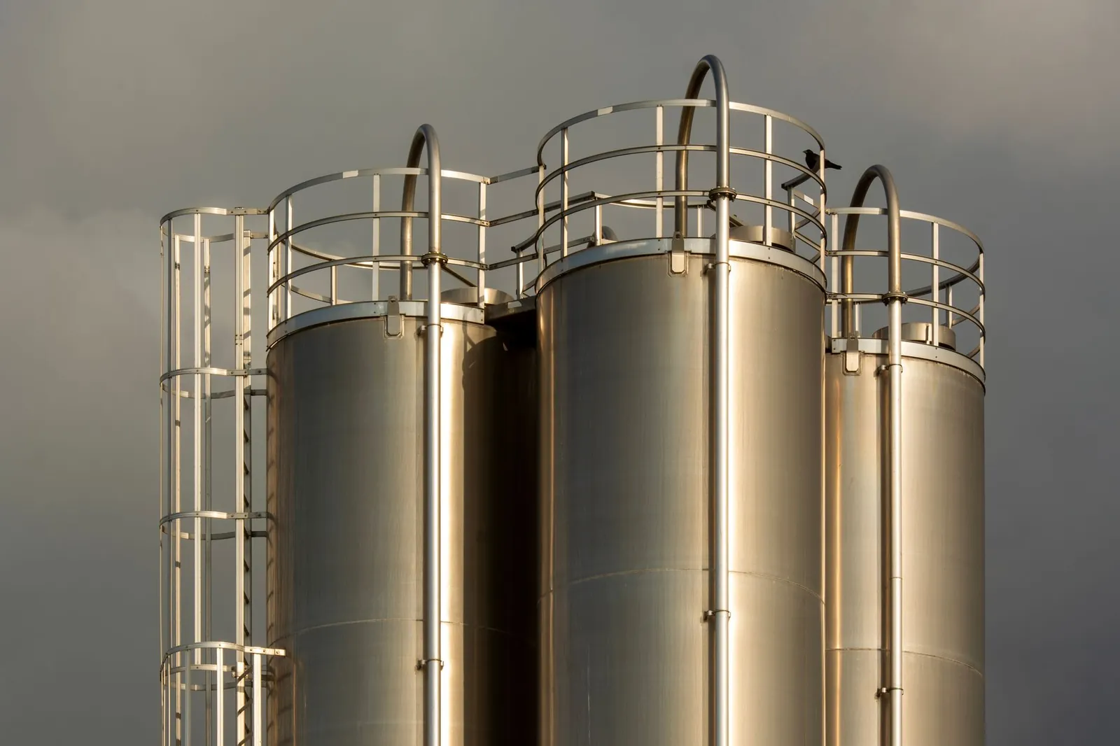 Close-up of industrial metal storage tanks against a backdrop of dark clouds, creating a dramatic industrial scene. Stock photo illustrating Laxmi Organic Industries' production started story.