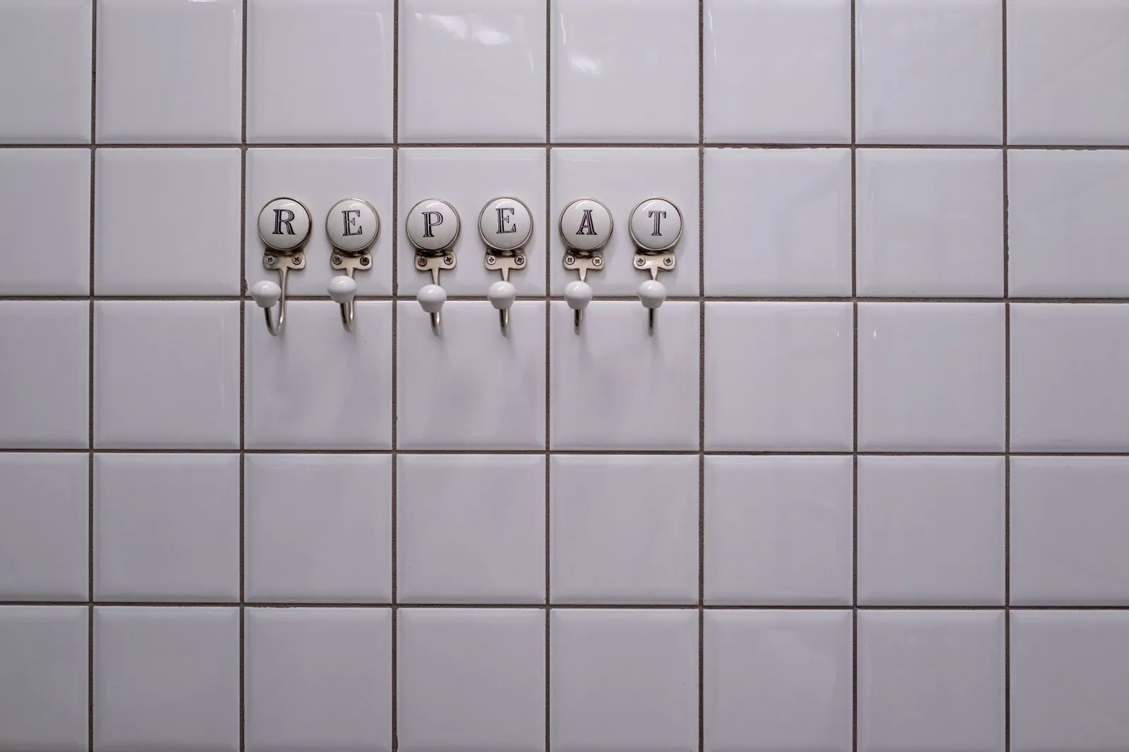 White tiled wall with towel hooks spelling 'REPEAT', showcasing a conceptual minimalist design. Stock photo for the ALFA News guide: Why Repeat Orders Matter for Indian Stock Investors.