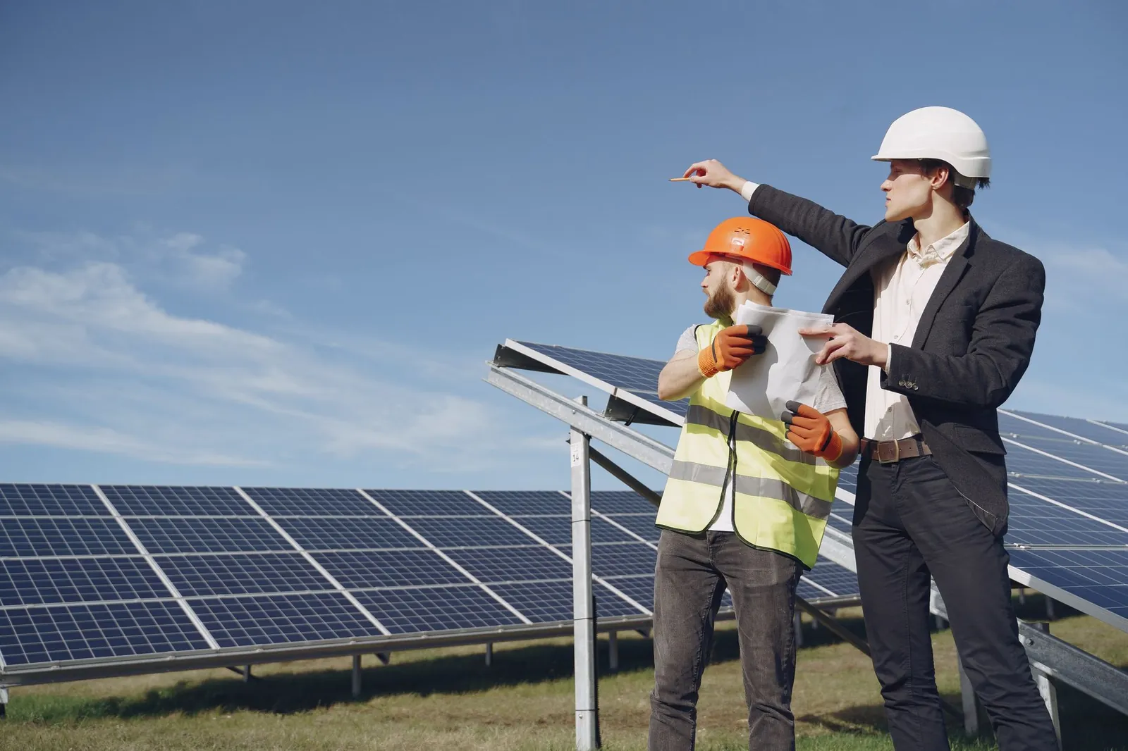 Young businessman in formal outfit and foreman in yellow vest looking away while discussing working process against solar panels. Stock photo illustrating Samvardhana Motherson International's production started story.