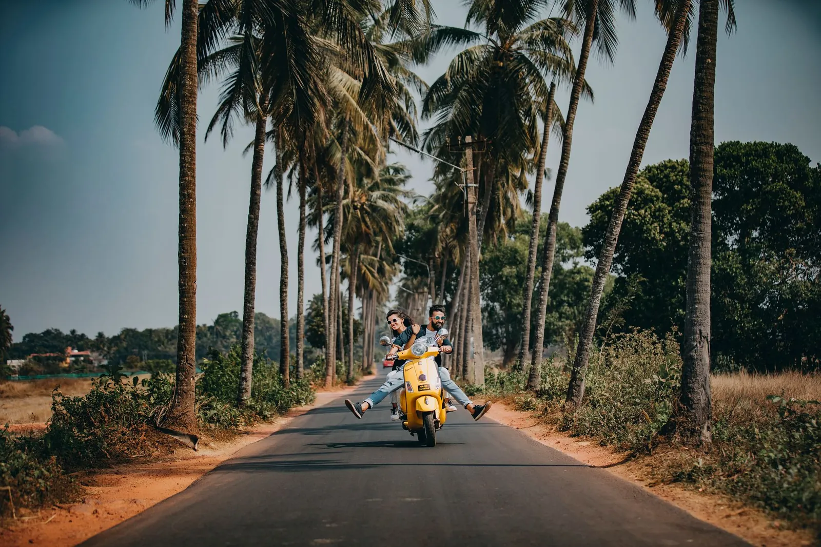 A joyful couple rides a yellow scooter through a scenic palm-lined road, capturing the essence of tropical travel and adventure. Stock photo illustrating Zelio E-Mobility's production started story.