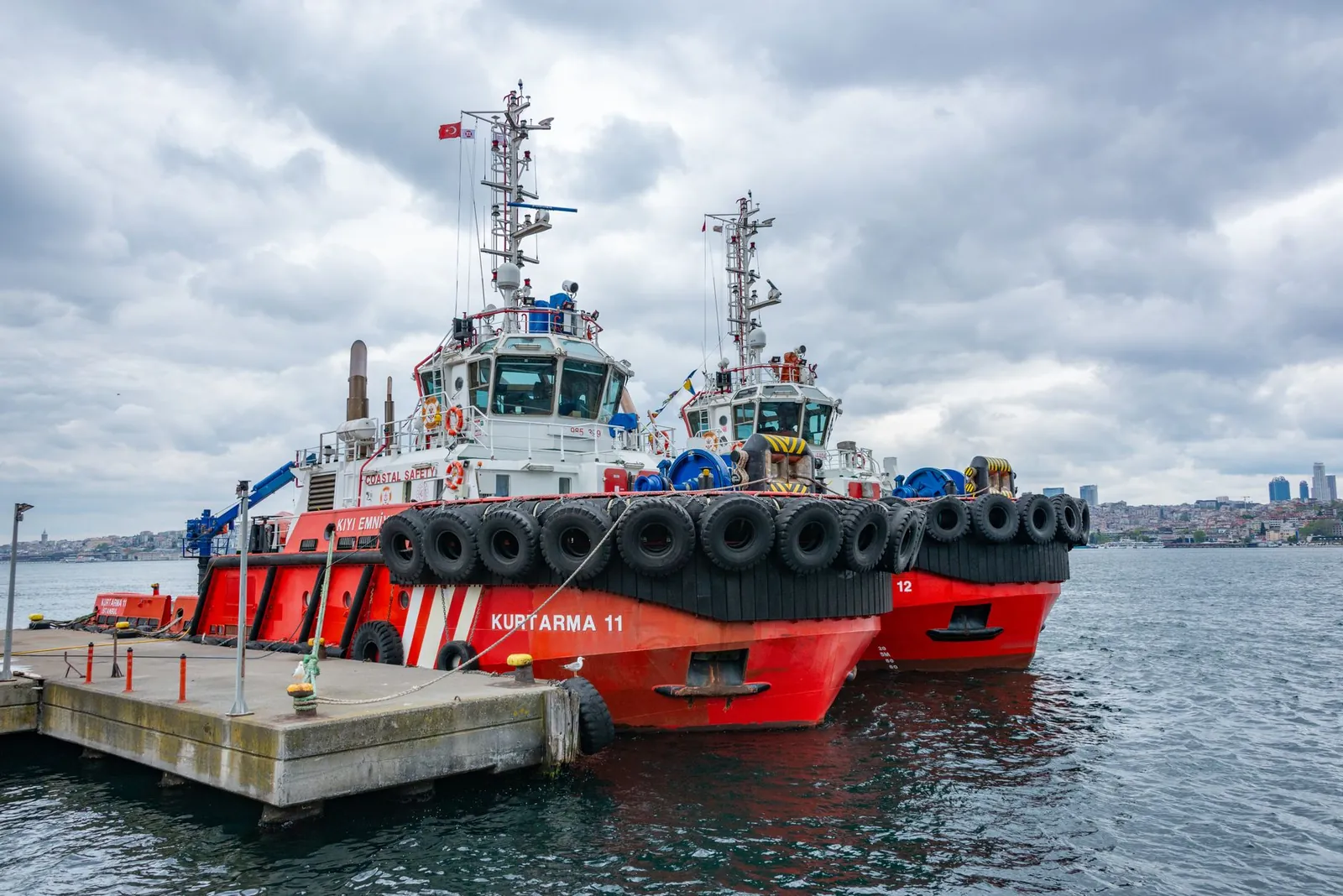 Two red tugboats docked under a cloudy sky at an urban harbor, ready for action. Stock photo illustrating Swan Defence and Heavy Industries' new order story.