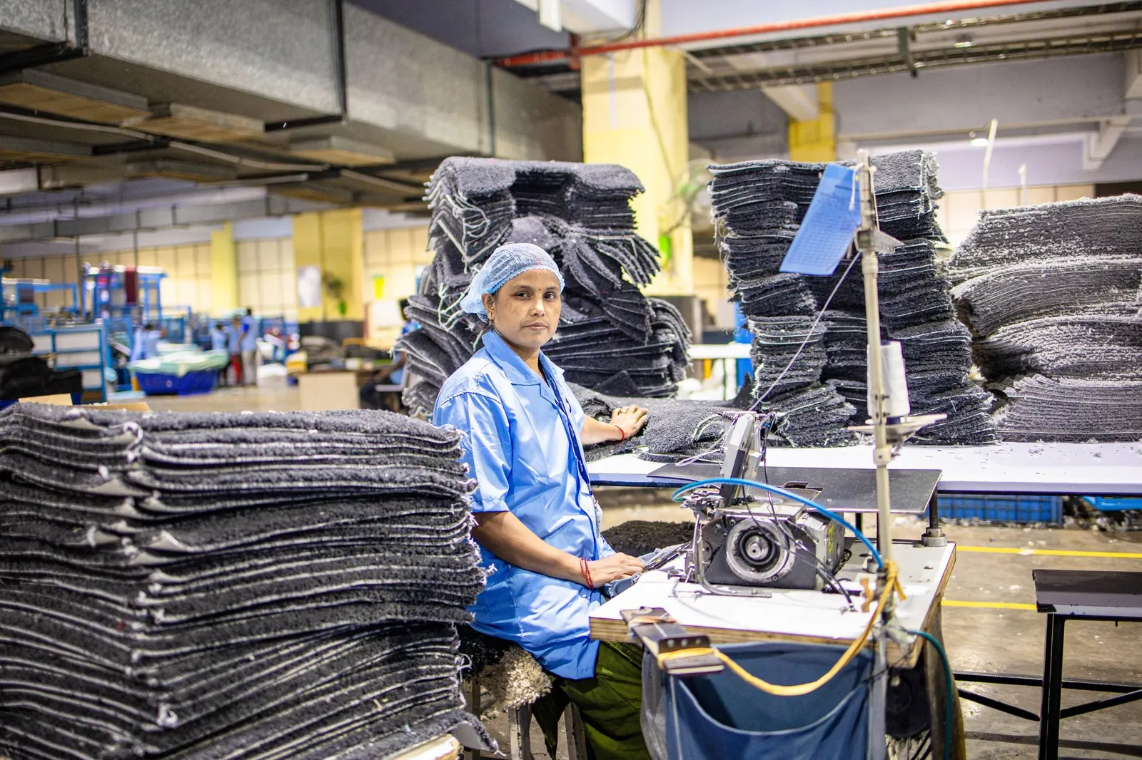 A factory worker operates a sewing machine surrounded by textile stacks in an industrial setting. Stock photo illustrating ACS Technologies' fund raise story.