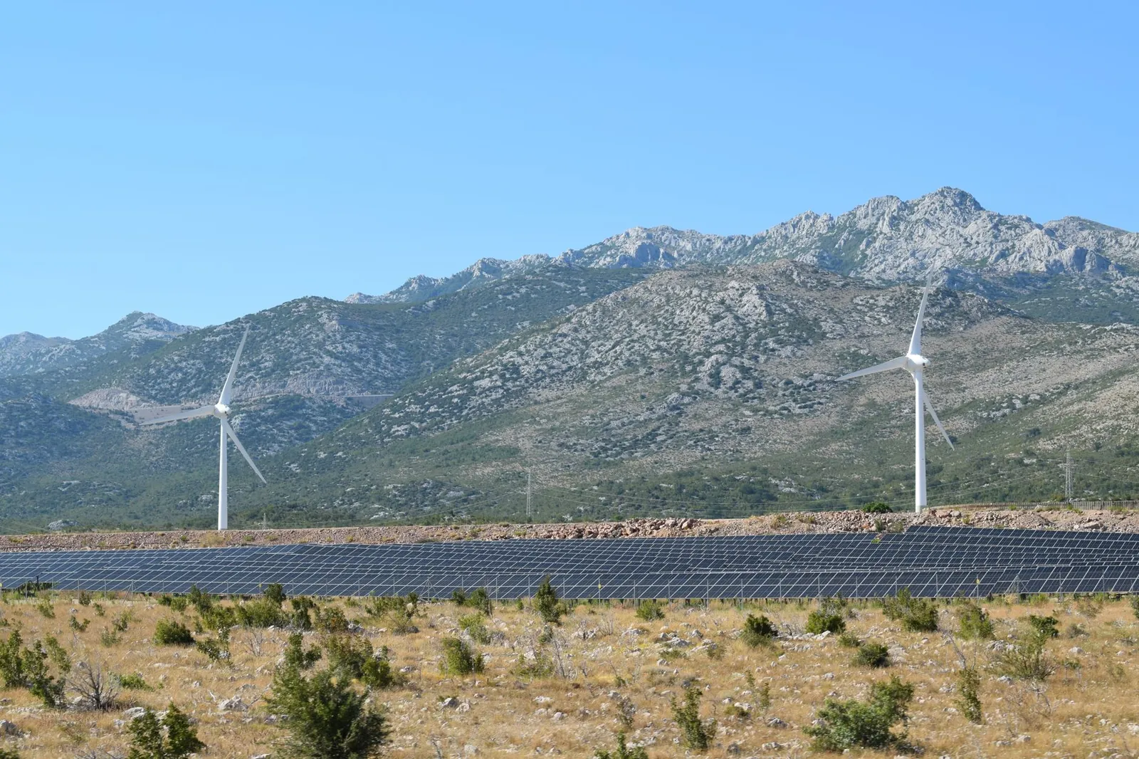 A scenic view of windmills and solar panels against mountainous terrain under a clear blue sky. Stock photo illustrating Alpex Solar's production started story.