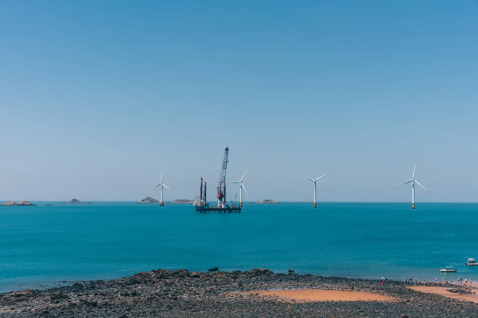 A serene view of offshore wind turbines against a clear blue sky with calm ocean waters. Stock photo illustrating Inox Wind's new order story.