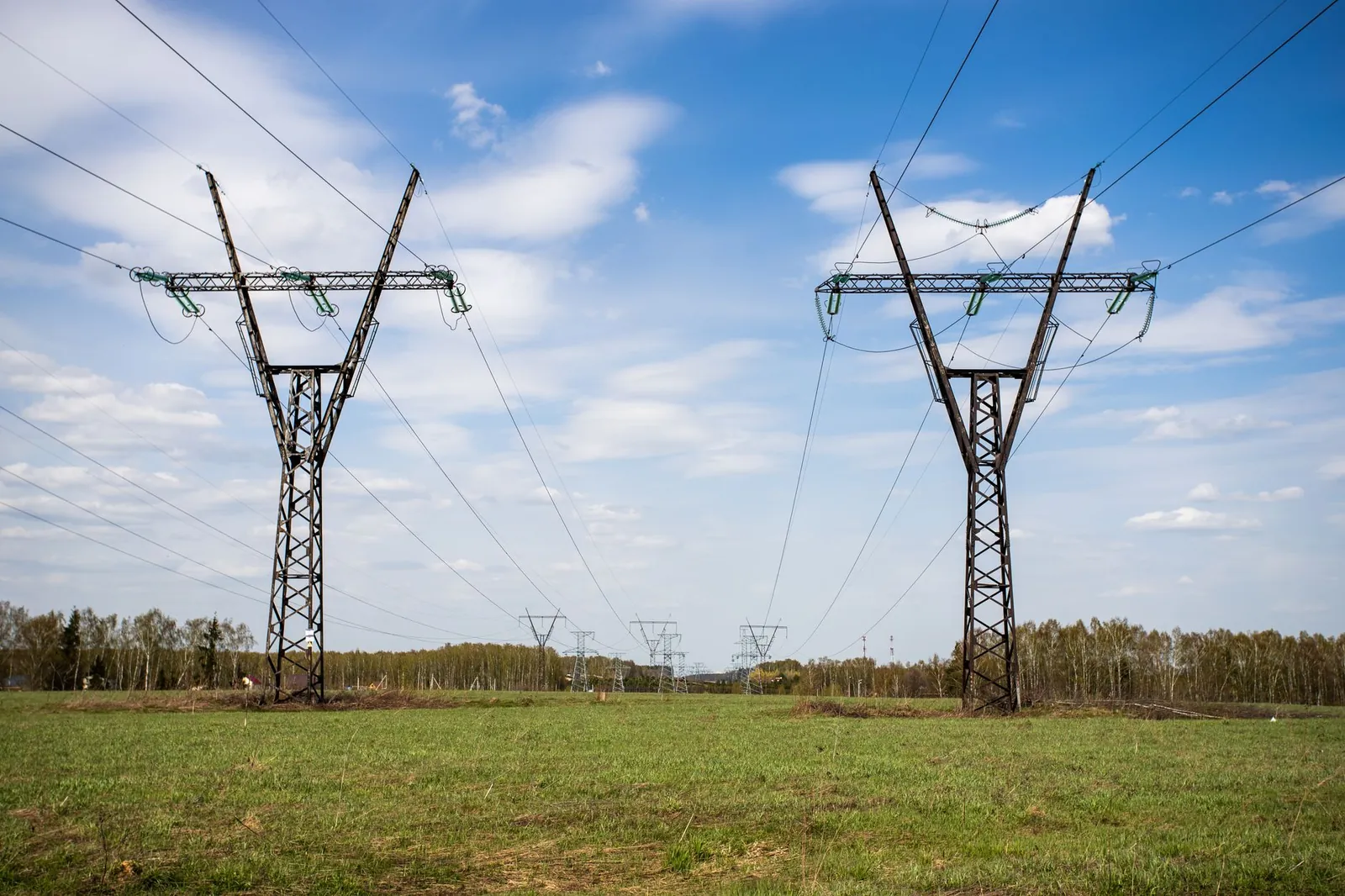 High voltage power lines stretch across a green grass field under a clear blue sky. Stock photo illustrating HEC Infra Projects' new order story.
