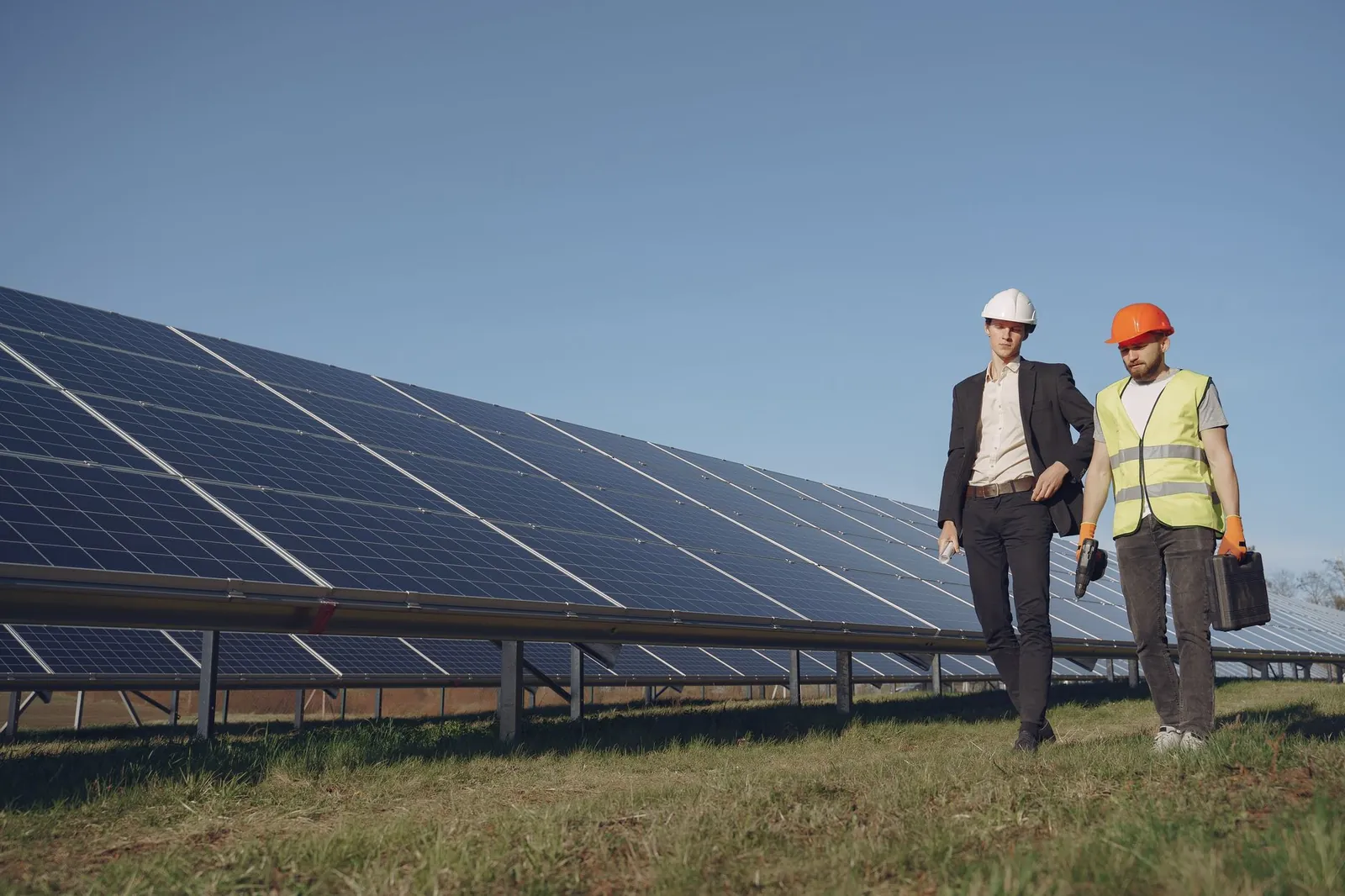 Full body of young focused manager in formal wear and electrical technician in uniform walking on path near solar panels and discussing working process. Stock photo illustrating Insolation Energy's new order story.