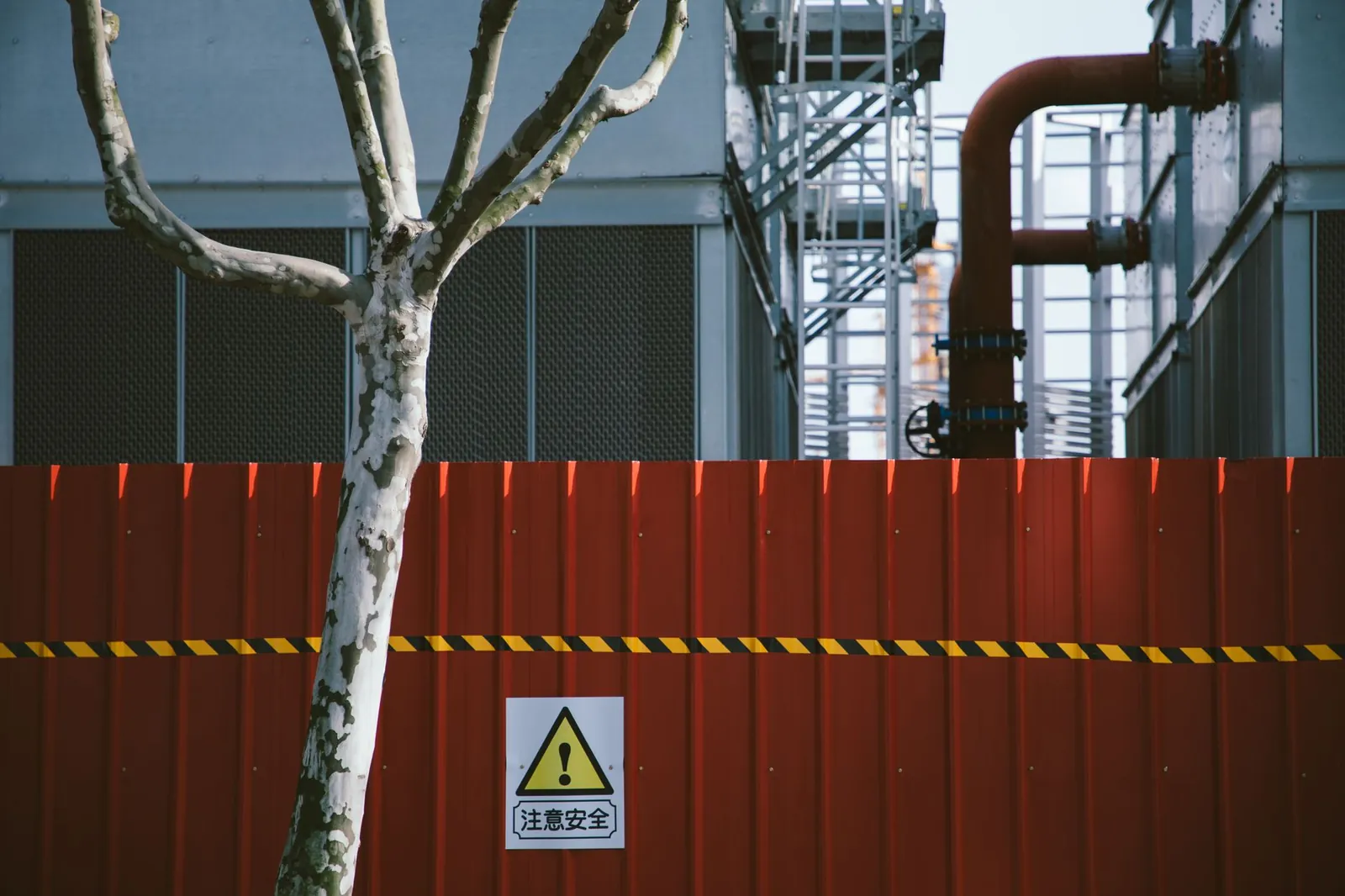 A red fence with a warning sign at an industrial site, with a bare tree and pipe structures. Stock photo illustrating Tatva Chintan Pharma Chem's expansion story.