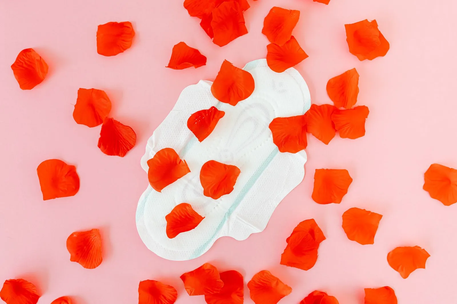 White sanitary napkin surrounded by red petals on a pink background, representing menstruation and feminine hygiene. Stock photo illustrating Amrutanjan Health Care's production started story.