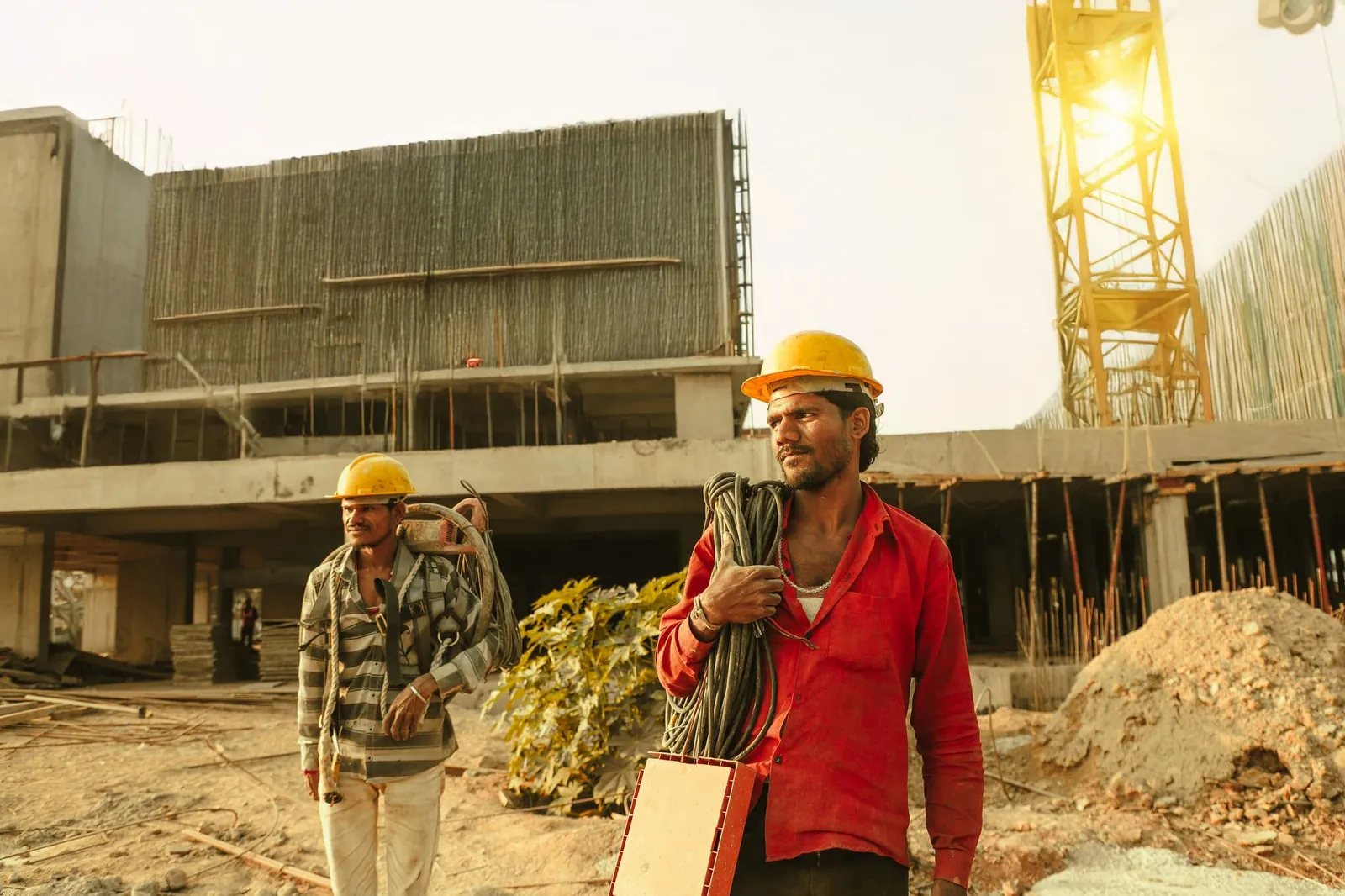 Two construction workers wearing helmets at a site in India with a crane and building. Stock photo illustrating Sathlokhar Synergys E&C Global's new order story.