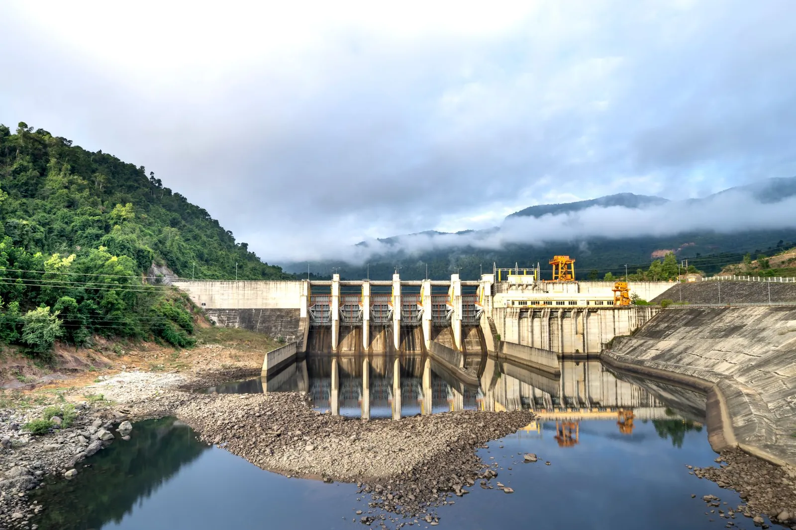 Industrial hydroelectric power plant located near river and stony coast against green mountain range covered with mist on summer day. Stock photo illustrating SJVN's financial results story.