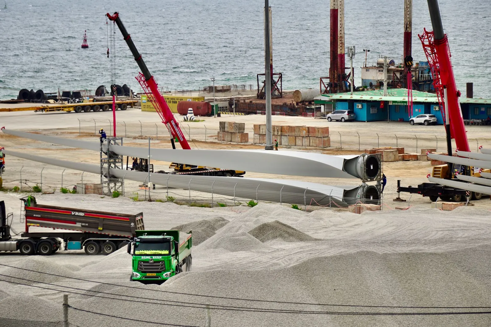 Construction site featuring wind turbine blades, cranes, and trucks by the seaside, showcasing industrial activity. Stock photo illustrating Enviro Infra Engineers' new order story.