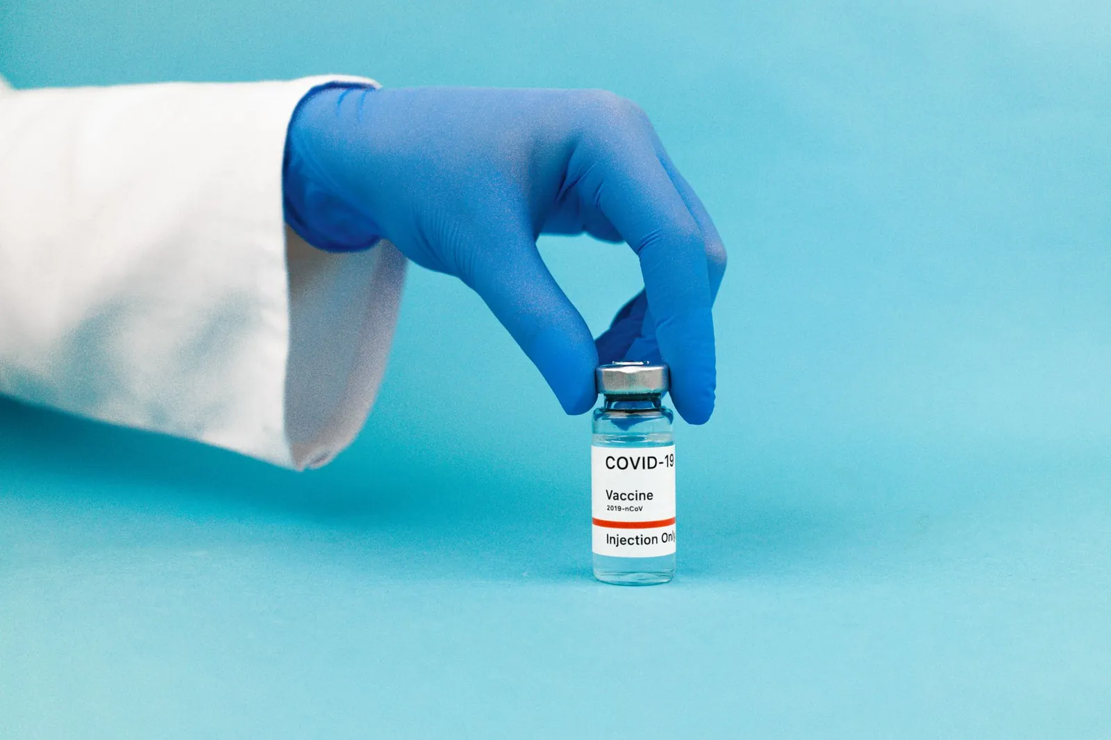 Medical professional hand wearing glove holding COVID-19 vaccine vial against blue background. Stock photo illustrating Biocon's regulatory approval story.