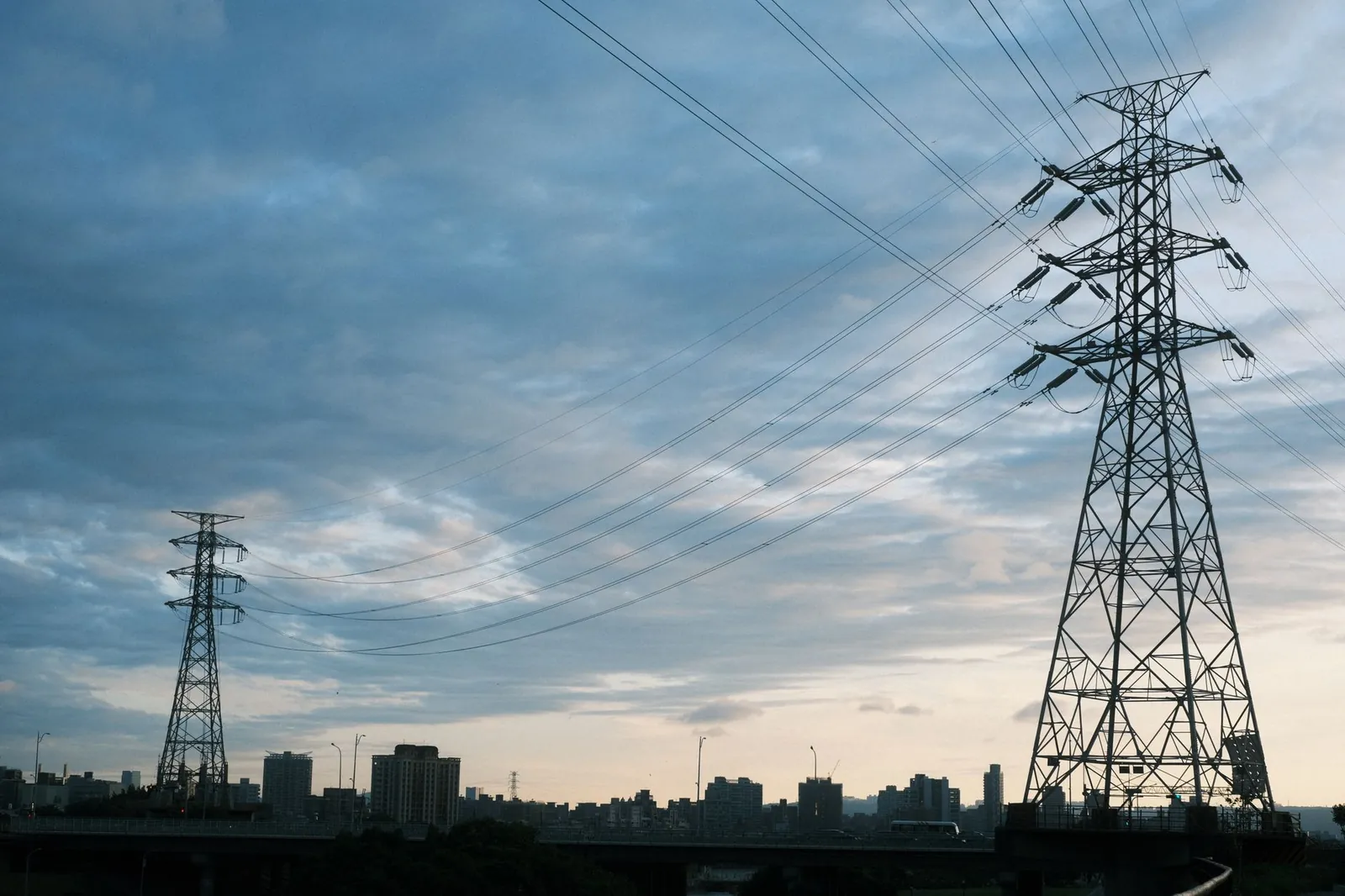 A city skyline at dusk with transmission towers and power lines silhouetted against a cloudy sky. Stock photo illustrating Transrail Lighting's earning call story.