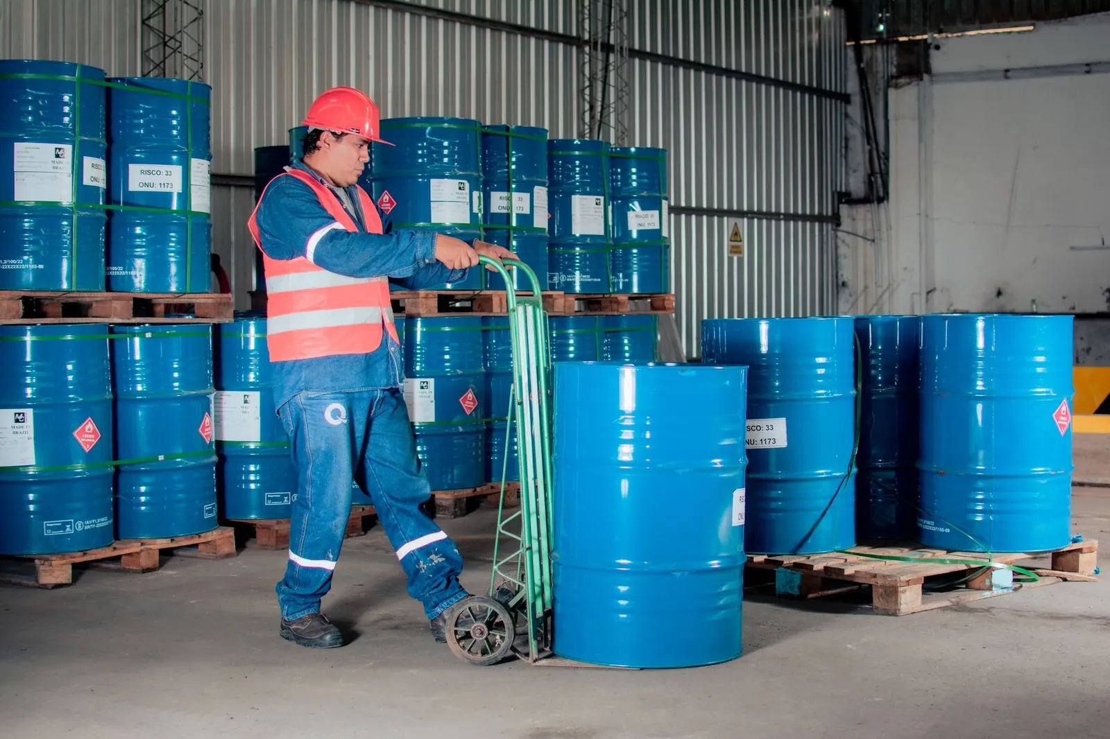 Industrial worker with a hand truck moving blue barrels in a warehouse setting. Stock photo illustrating Vishnu Chemicals' production started story.