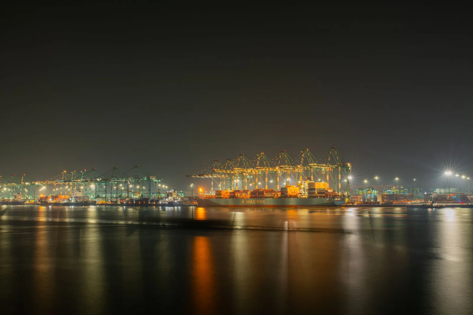 A serene nighttime view capturing the illuminated cranes and cargo ships at a bustling industrial shipping port. Stock photo illustrating Adani Ports and Special Economic Zone's financial results story.