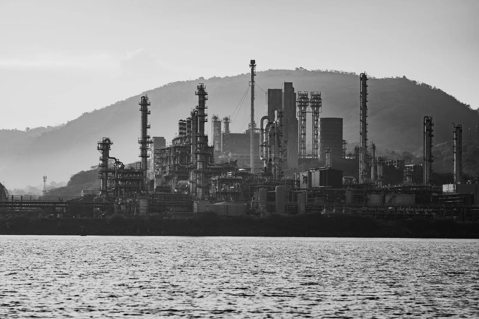 Black and white image of an industrial refinery set against a mountainous backdrop with water in the foreground. Stock photo illustrating Sadhana Nitro Chem's new order story.
