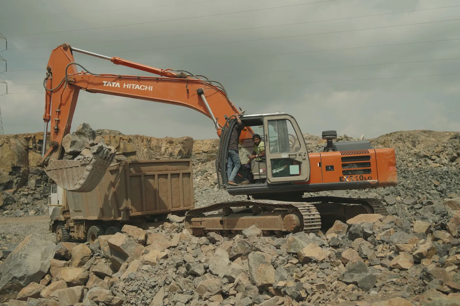 Excavator loading rocks into a dump truck at a quarry site, showcasing industrial equipment in action. Stock photo illustrating Tinna Rubber and Infrastructure's production started story.
