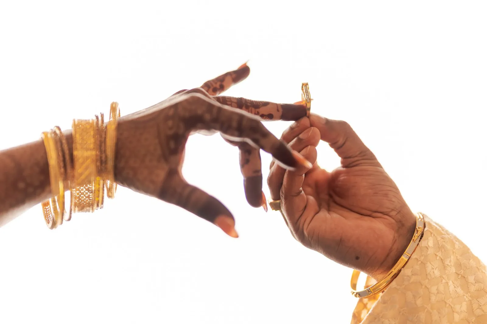 Close-up of hands exchanging rings, adorned with intricate henna and gold jewelry, in a traditional Indian wedding. Stock photo illustrating Motisons Jewellers' fund raise story.