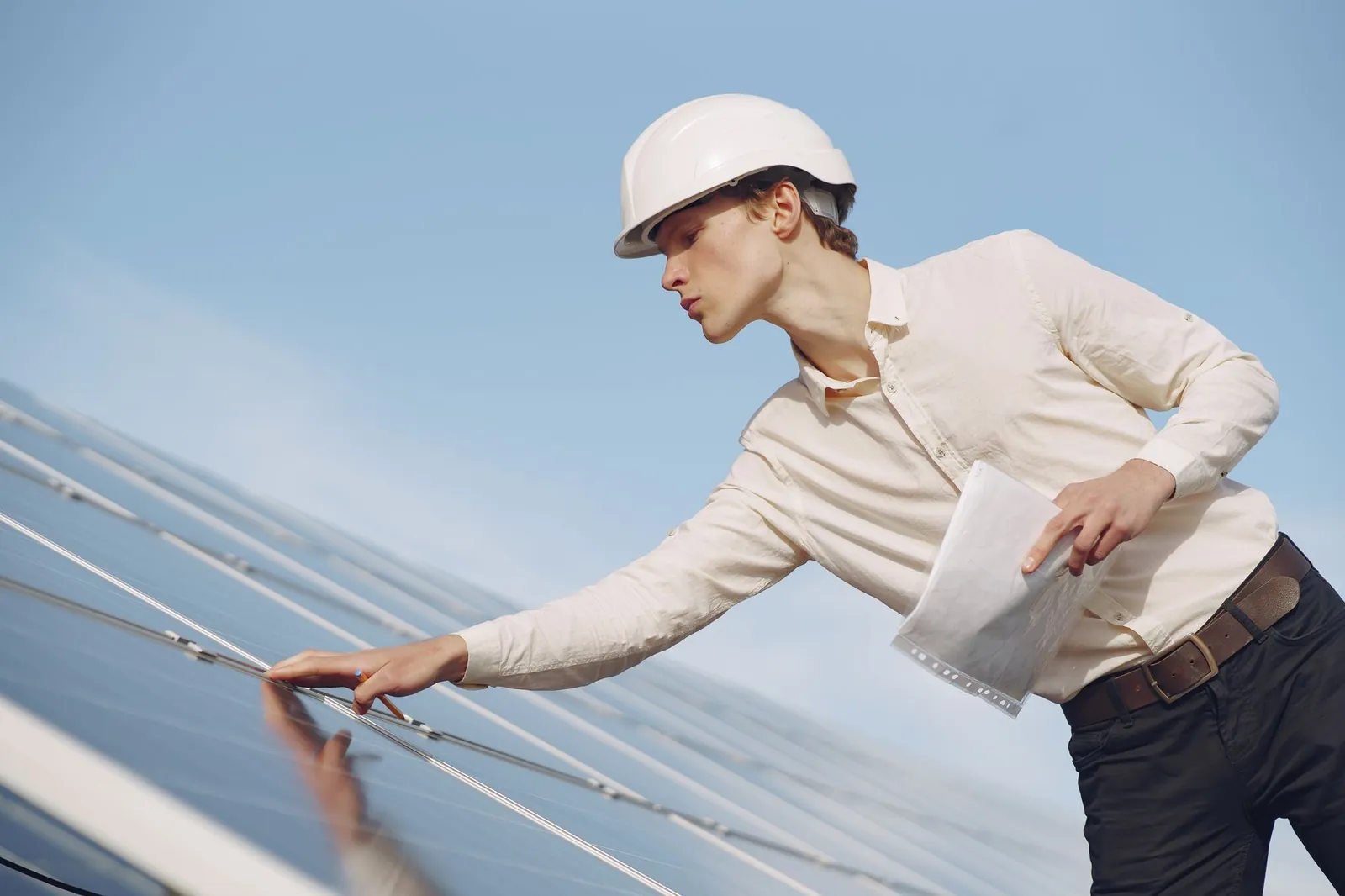 Low angle of young male inspector with documents in formal wear and hardhat standing against solar panels and checking details of construction. Stock photo illustrating Advait Energy Transitions' new order story.