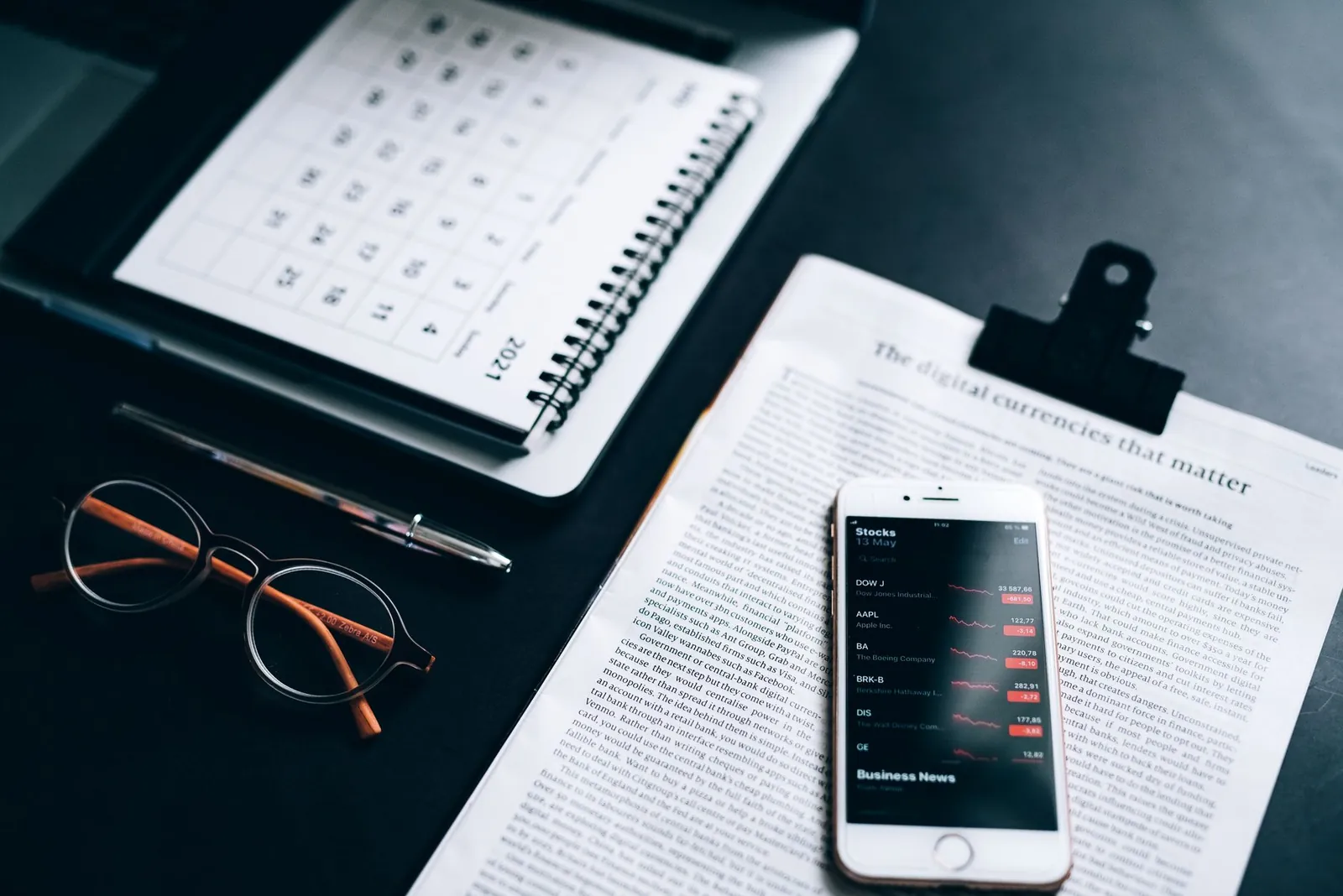 High angle shot of a desk setup with smartphone displaying stock market analytics, clipboard, pen, and glasses. Stock photo for the ALFA News guide: How to Get Stock Market News Faster.